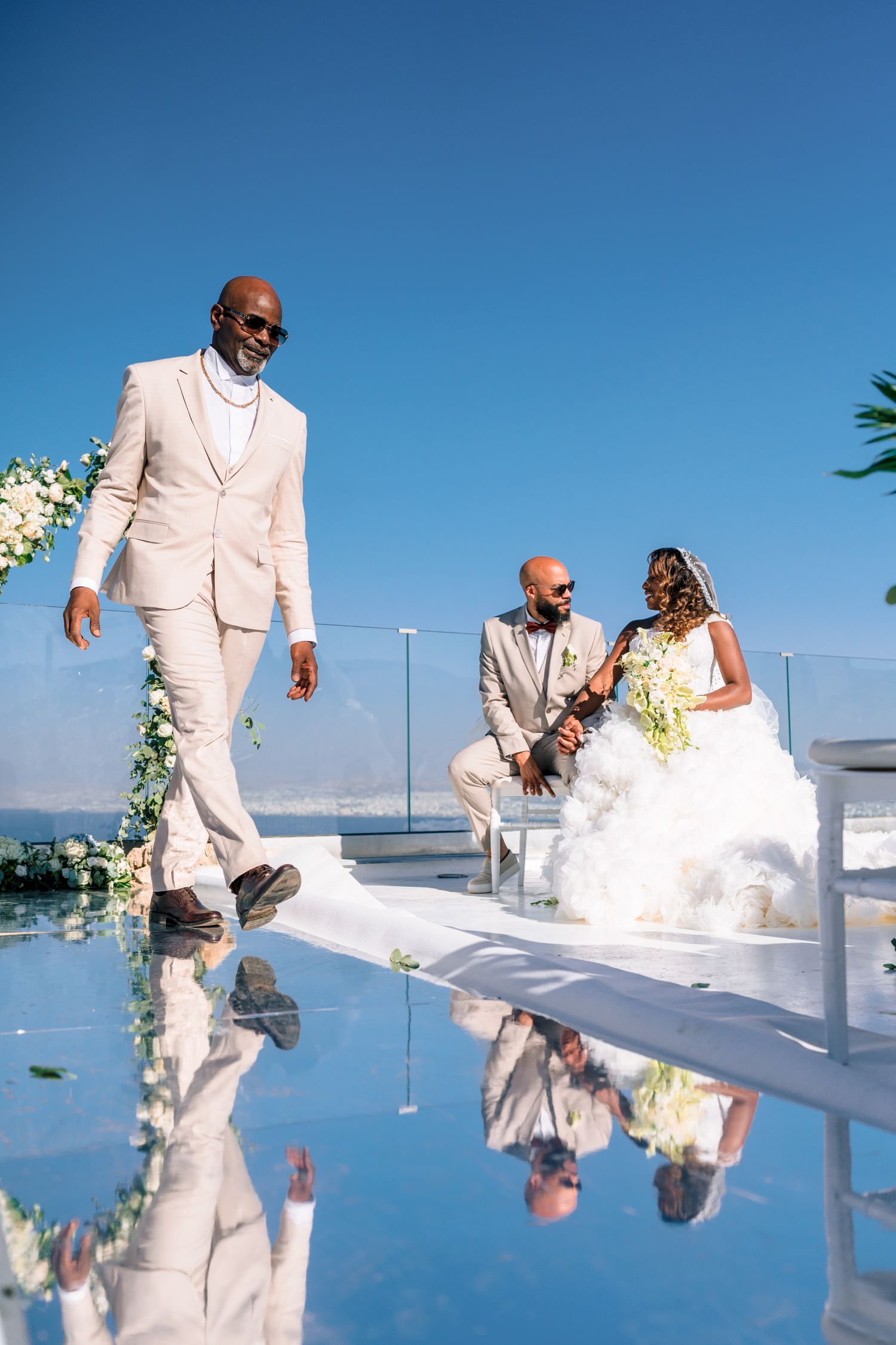 Wedding couple in white attire reflected in infinity pool against blue sky.