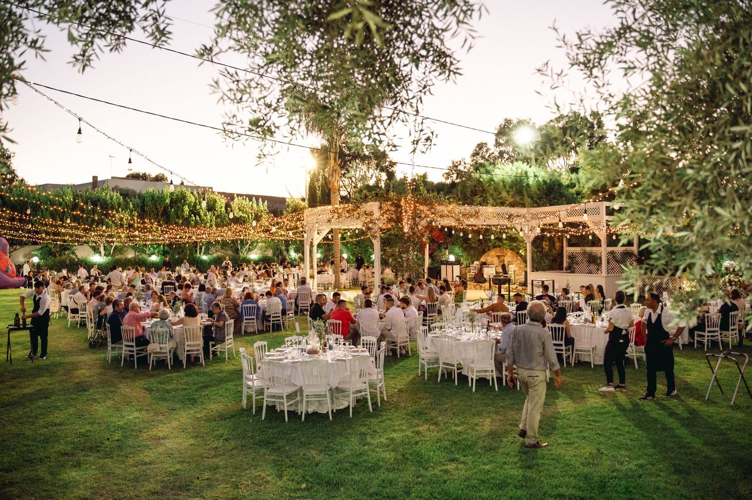 Panoramic view of an outdoor wedding reception with tables set under trees at sunset.
