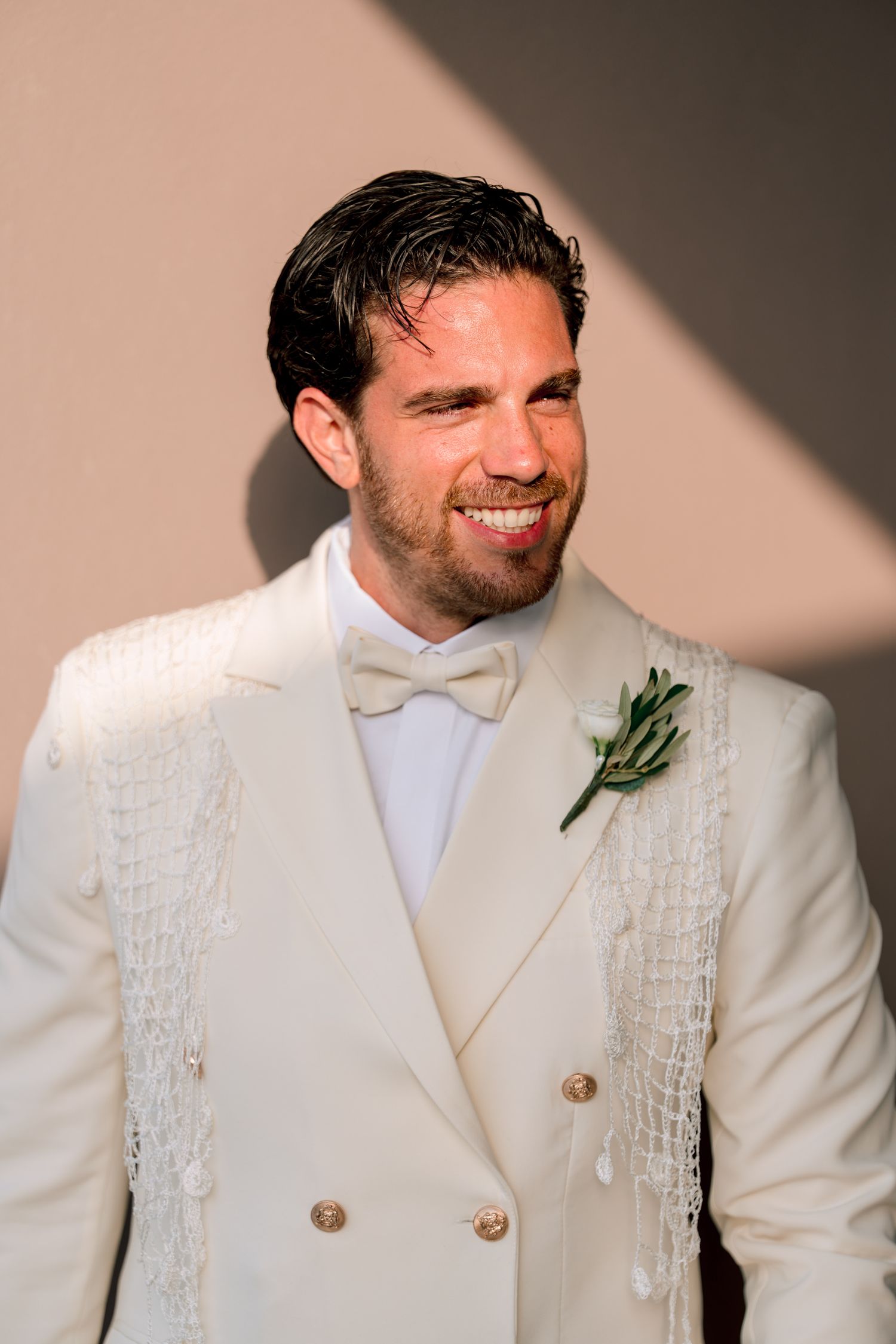Portrait of groom in cream double-breasted tuxedo with boutonniere against neutral background.