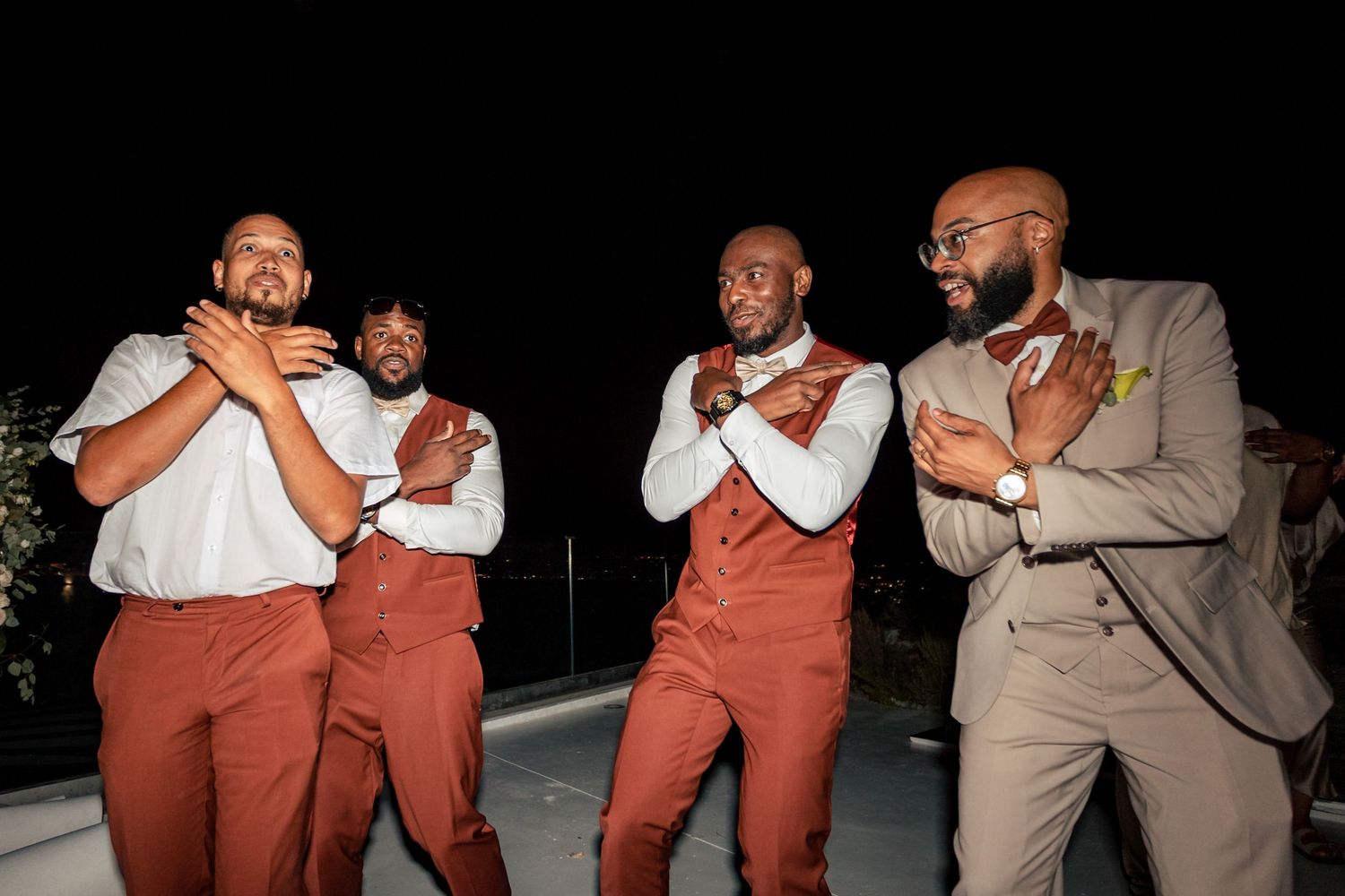 Groomsmen in rust-colored pants strike playful poses during wedding celebrations.