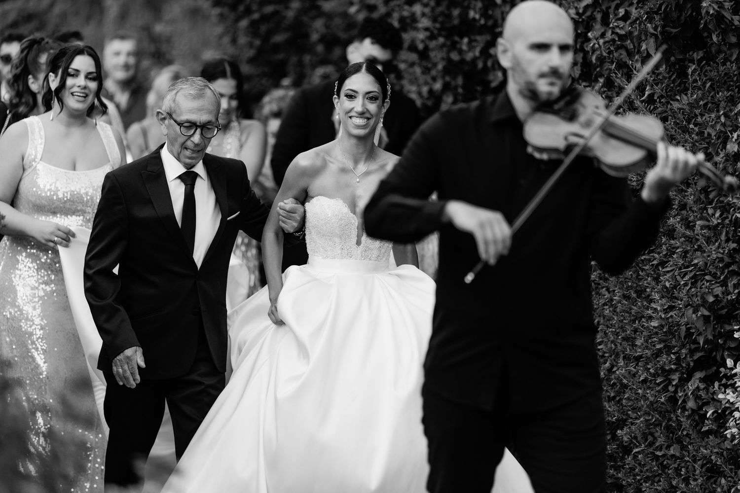 Wedding celebration procession with violinist leading through garden setting.