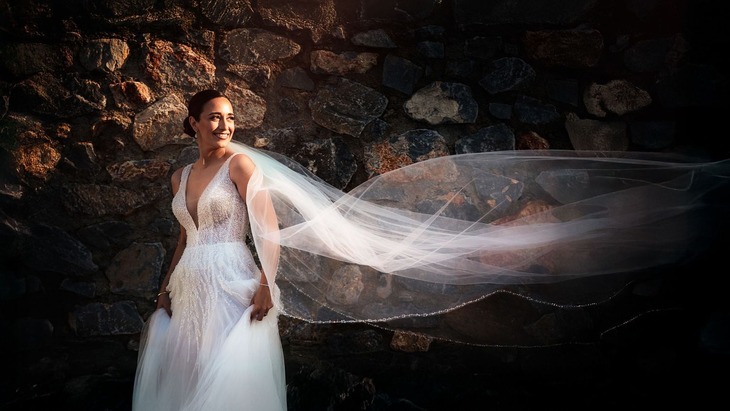 Dramatic bridal portrait with flowing veil against textured stone wall background.