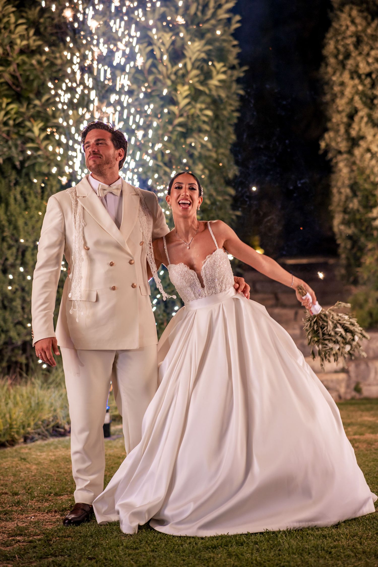 Joyful wedding couple in all white attire dance under sparklers at night.