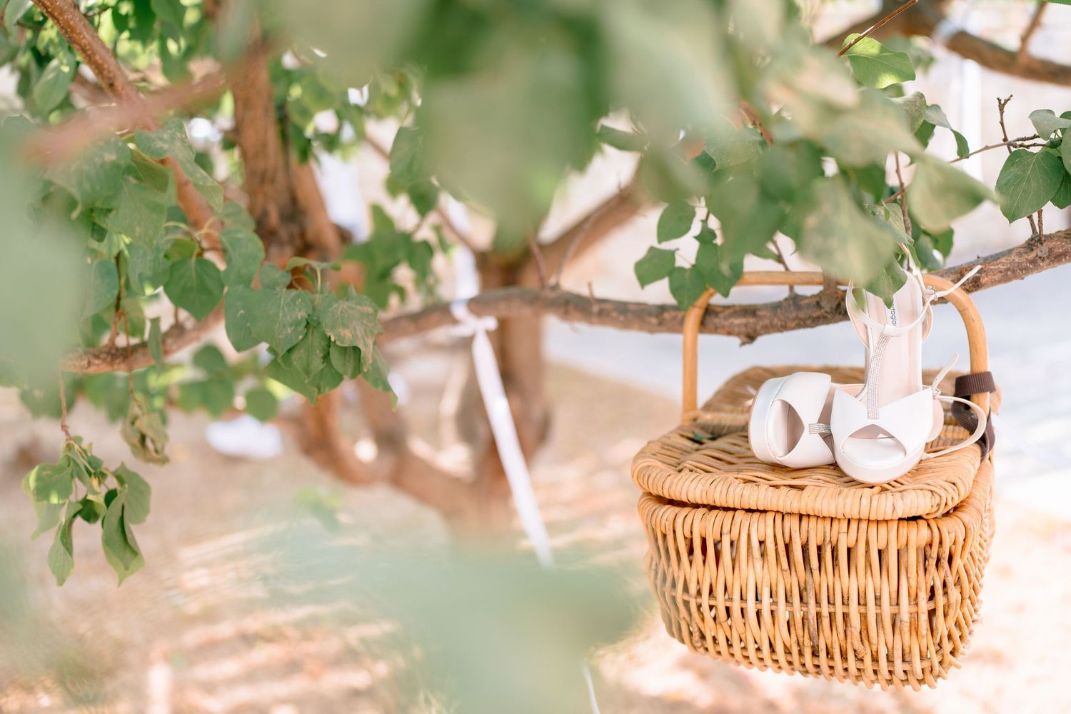Wicker basket with white shoes under green foliage at an outdoor wedding setting.