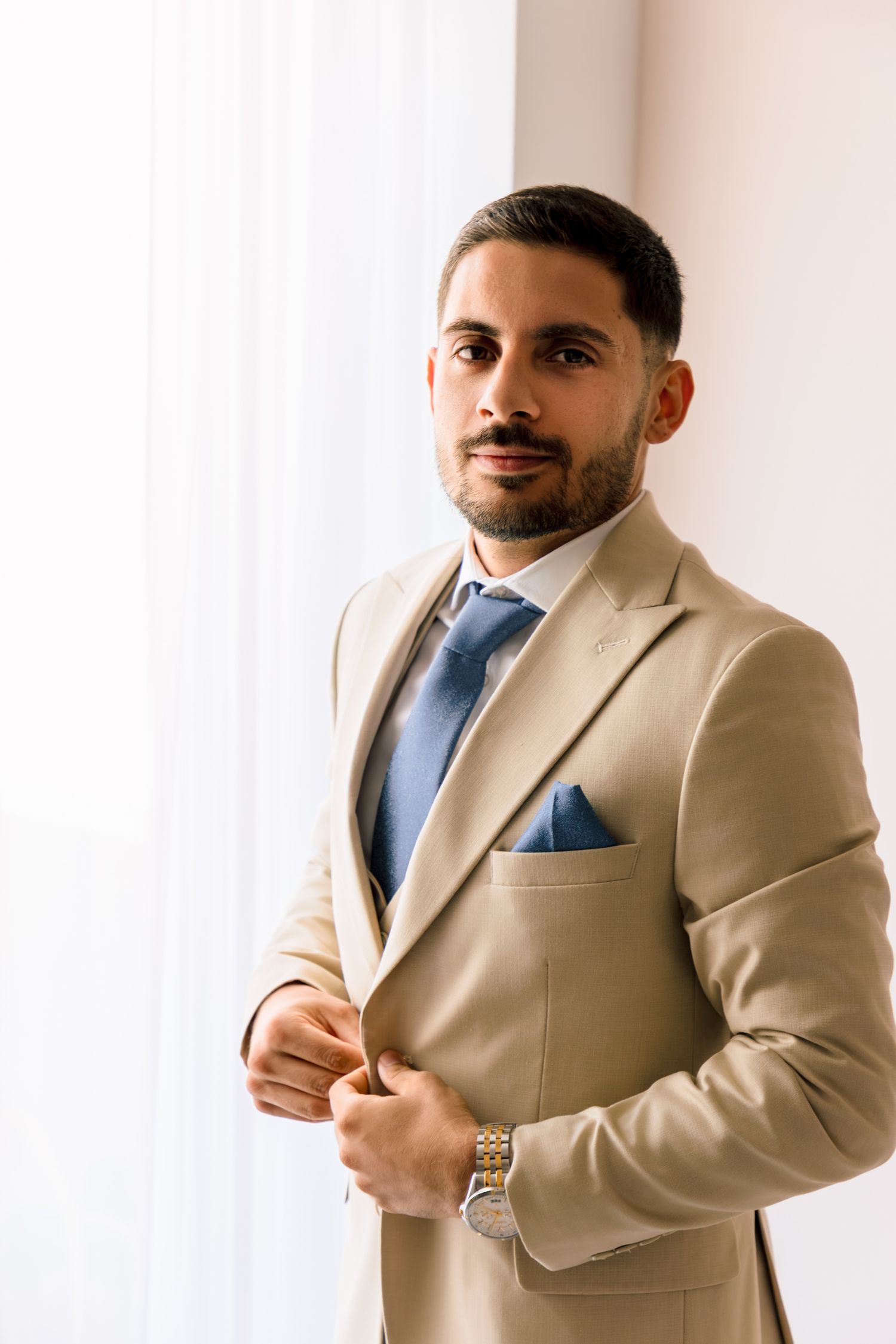 A person in a beige suit and blue tie poses confidently against a white wall.