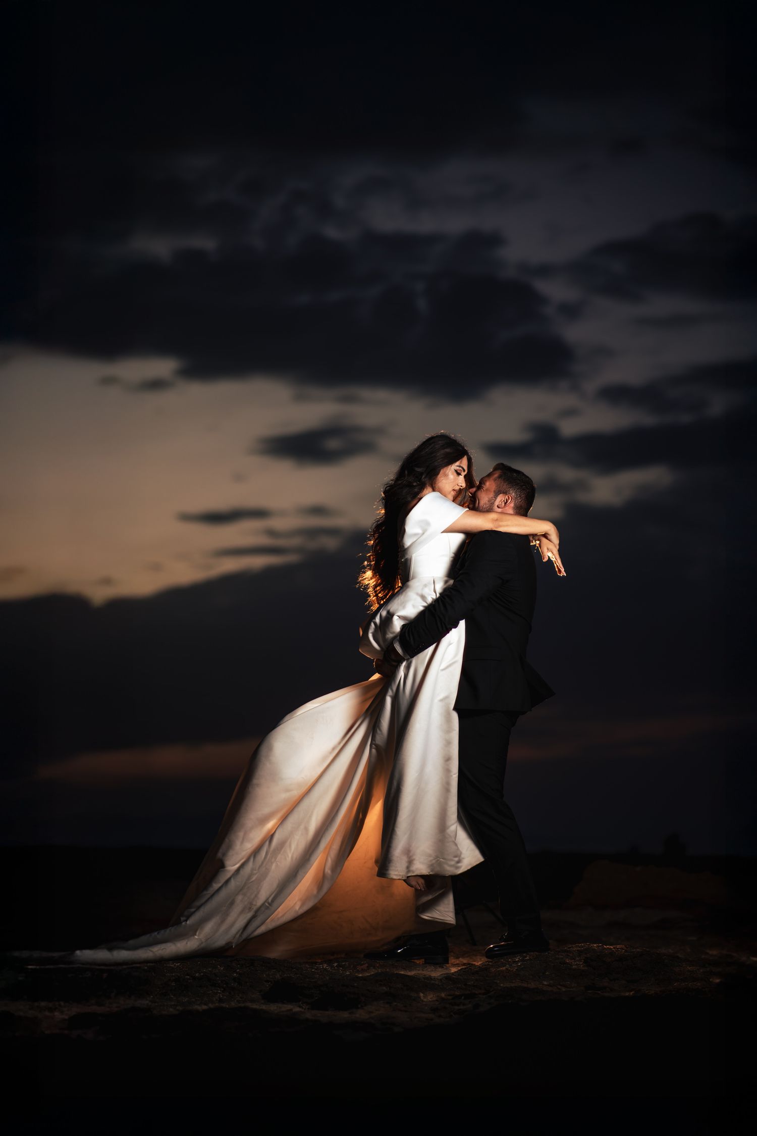 Dramatic sunset wedding photo with couple embracing against dark stormy sky.