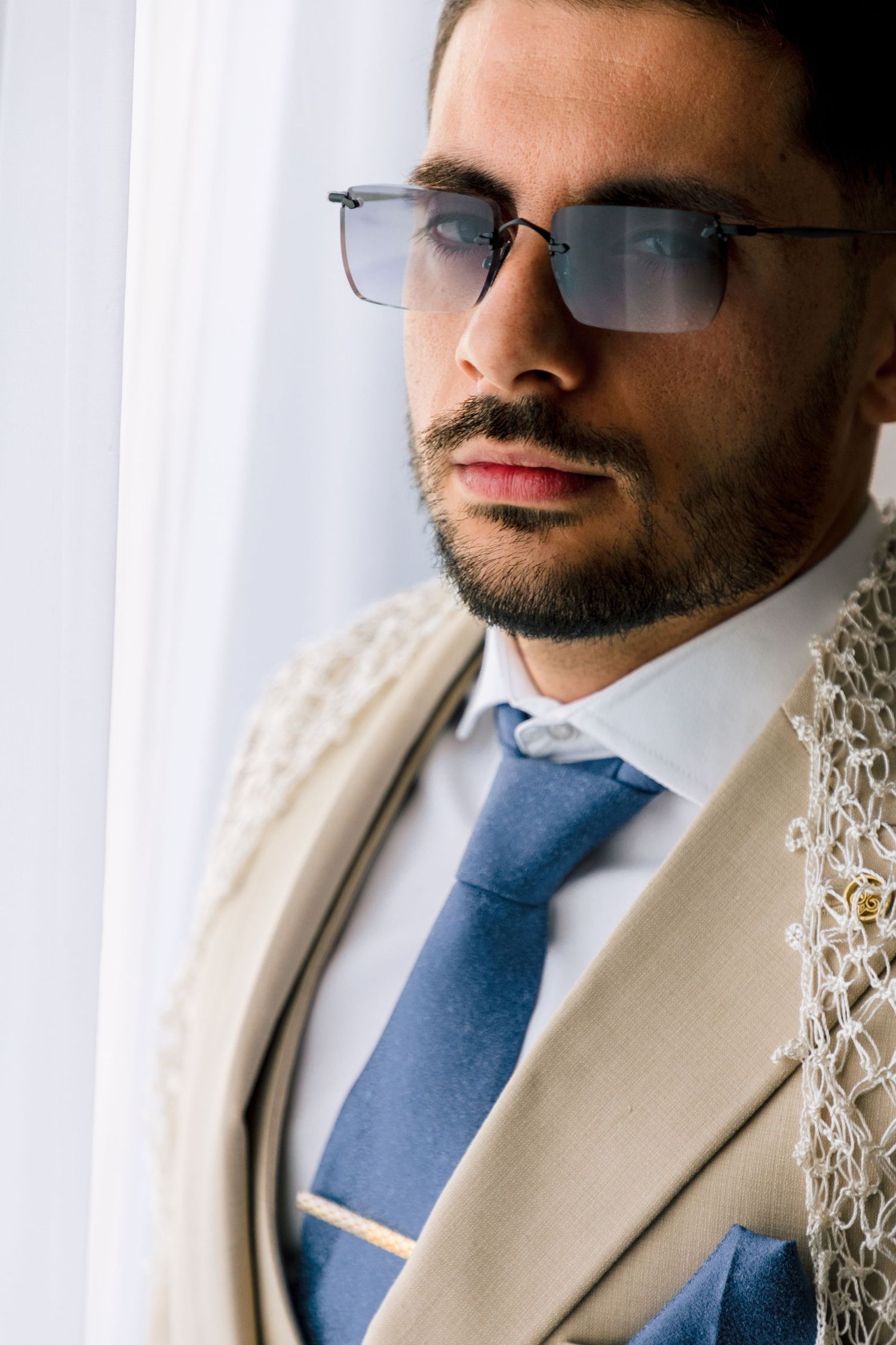 Person in beige suit and sunglasses with blue tie poses against white backdrop.