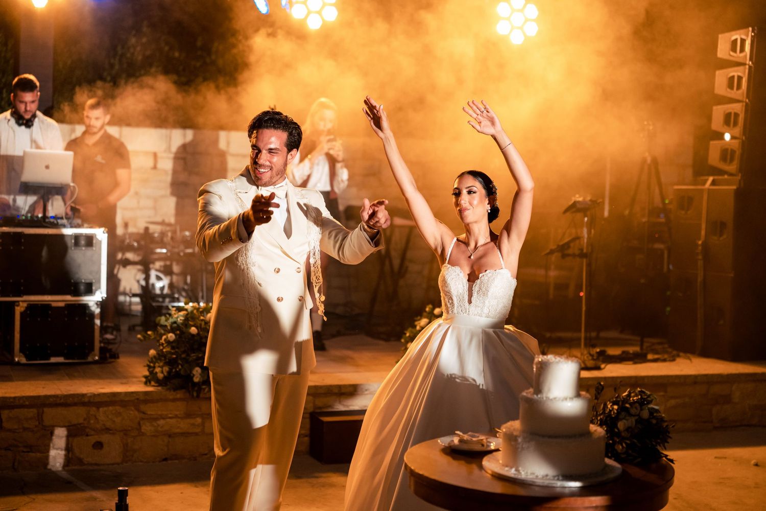 Newlyweds celebrate with joyful dance moves during their wedding reception under warm lighting and smoke effects.
