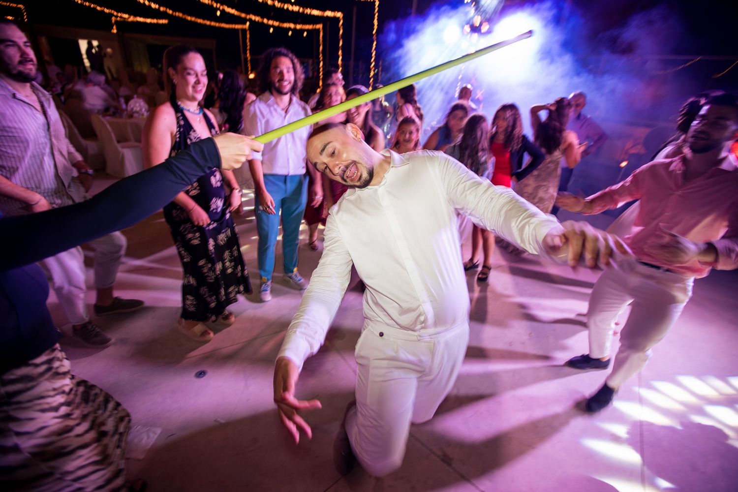 Dance floor illuminated with purple and blue lights as people dance beneath string lighting.