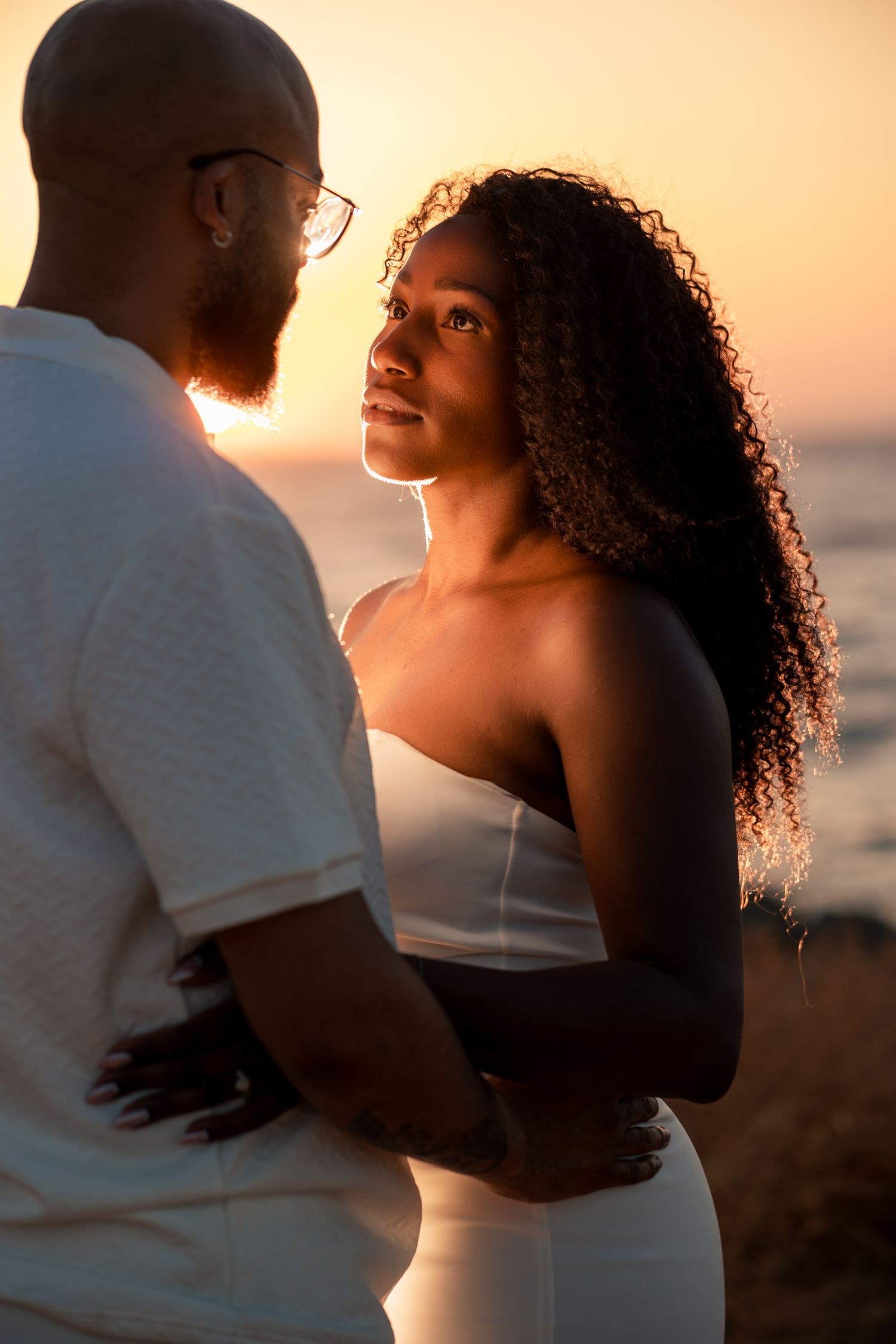 Couple shares tender moment at golden hour with ocean backdrop and warm lighting.