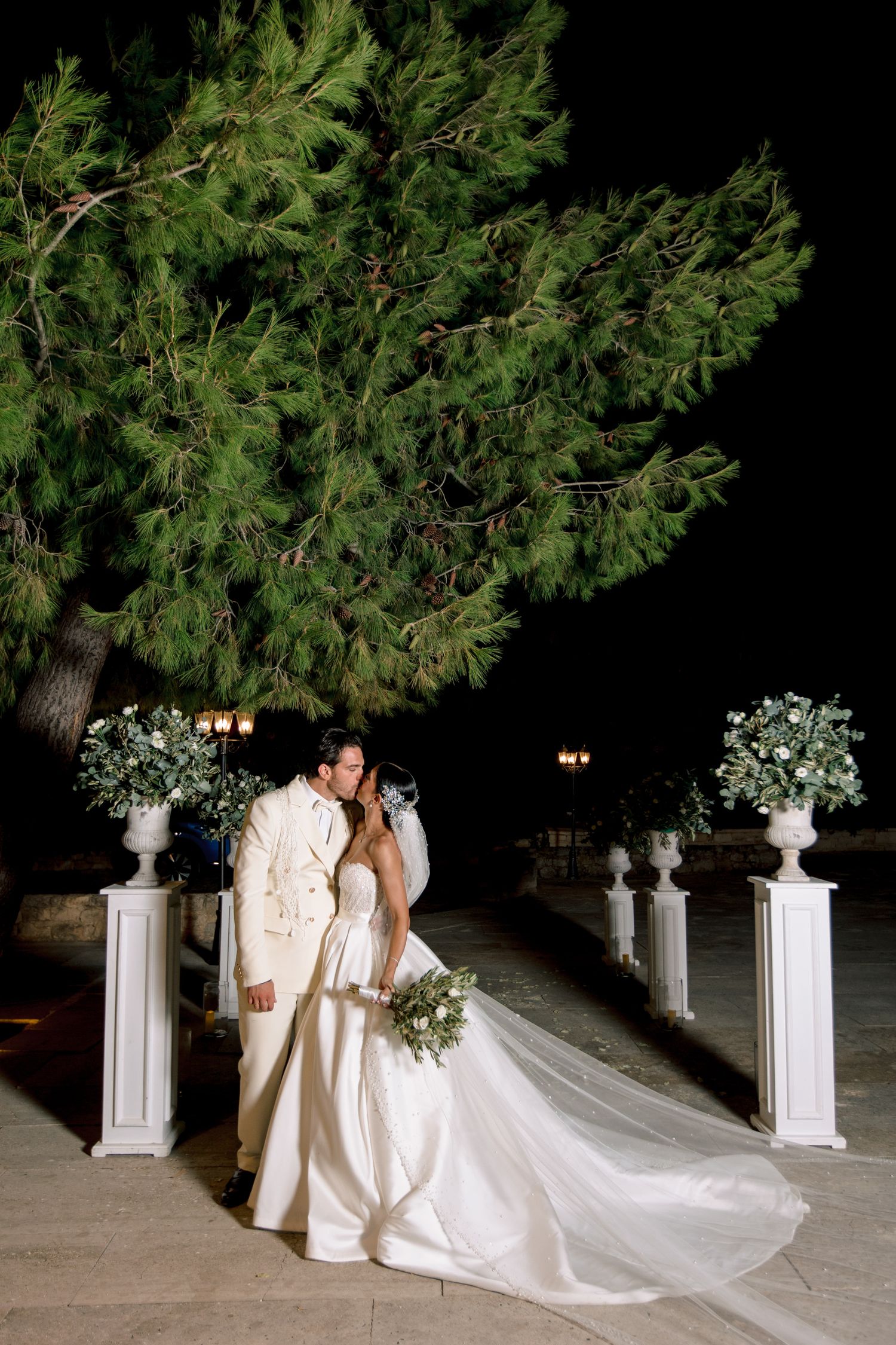 A romantic nighttime wedding portrait with white columns and evergreen trees in the background.