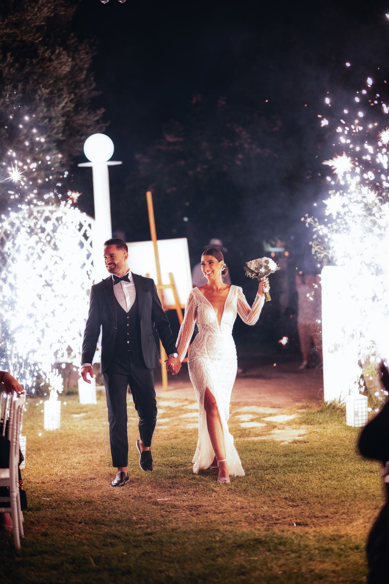 Sparklers illuminate a nighttime wedding celebration on a grassy field.