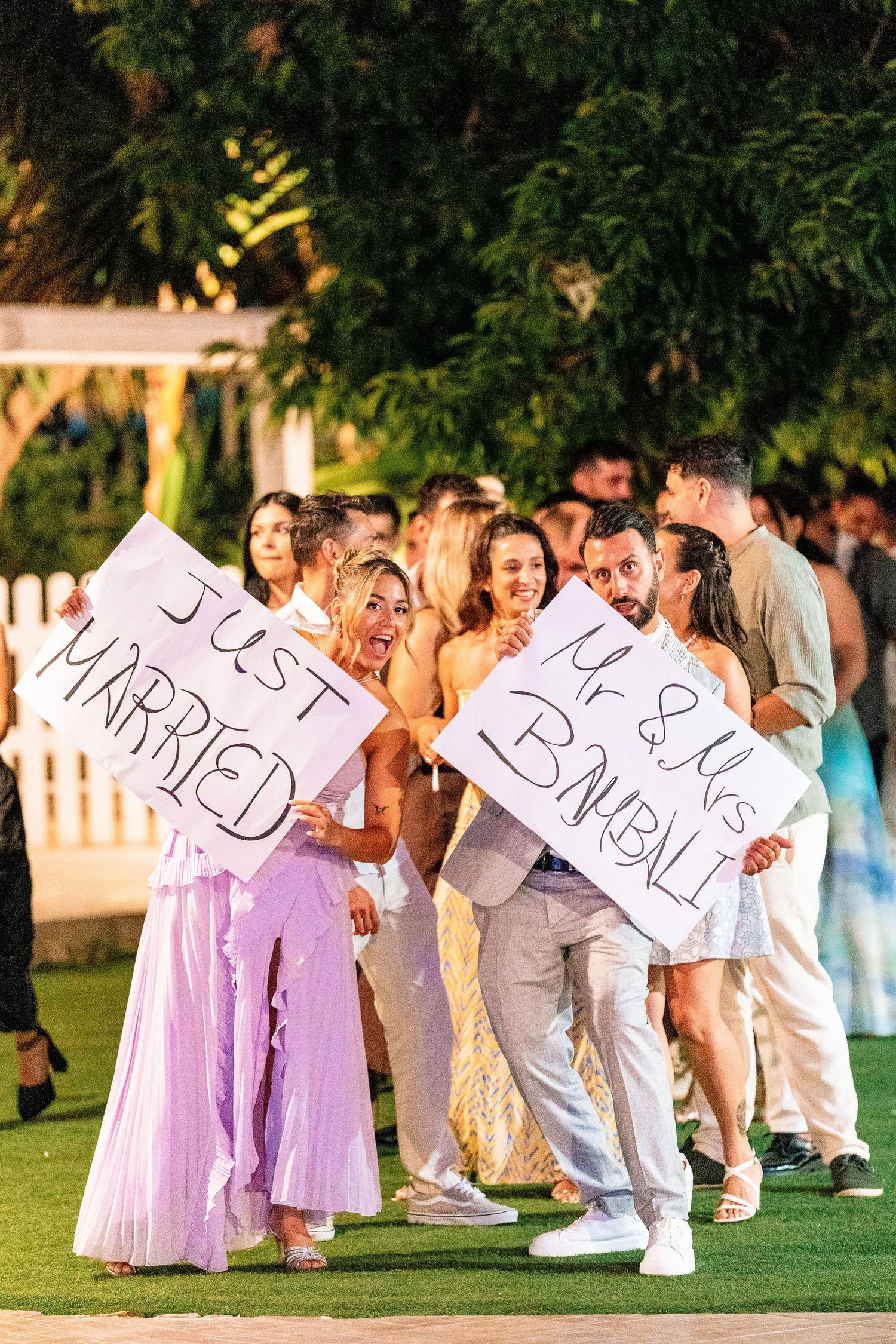 Bridal party in lavender dresses and white suits posing with fun signs on grass.