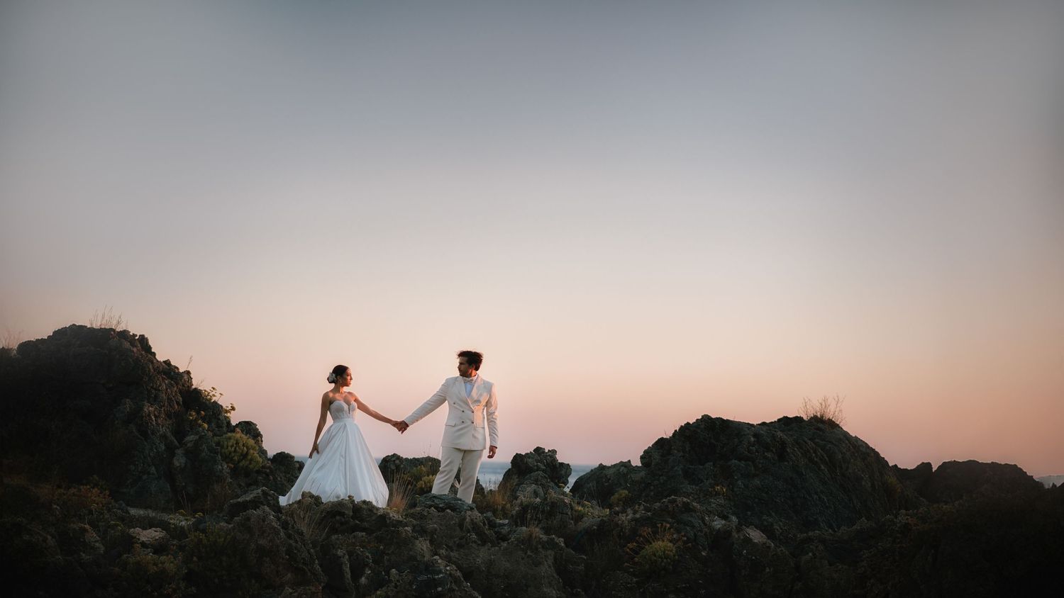 Couple in wedding attire embrace against dramatic rocky cliffs at sunset.