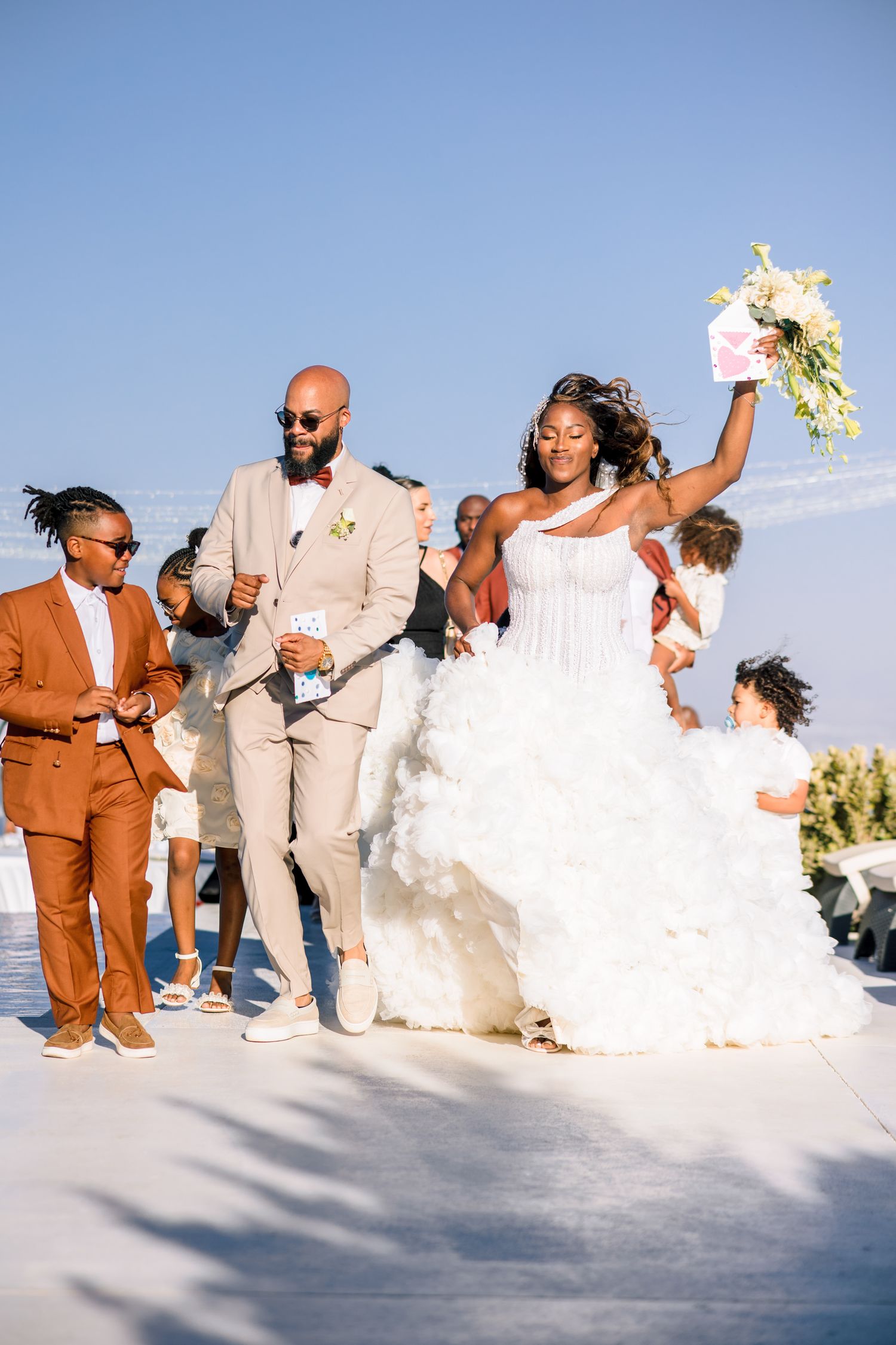 Wedding party poses on white surface against bright blue sky during outdoor celebration.