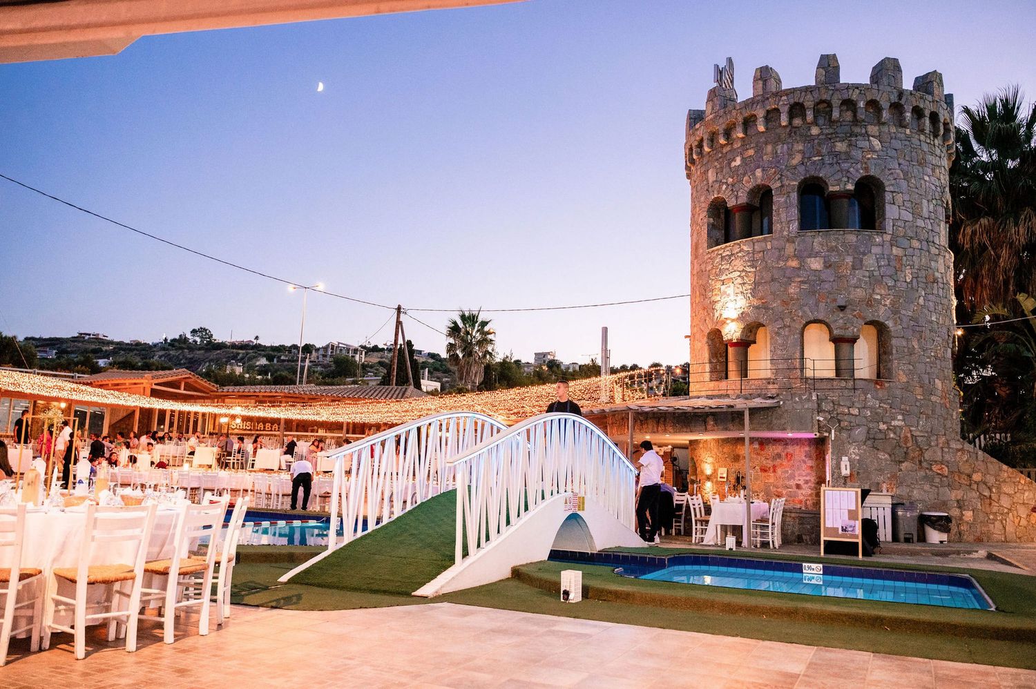 Wedding reception setup by a pool with stone castle tower and palm trees.