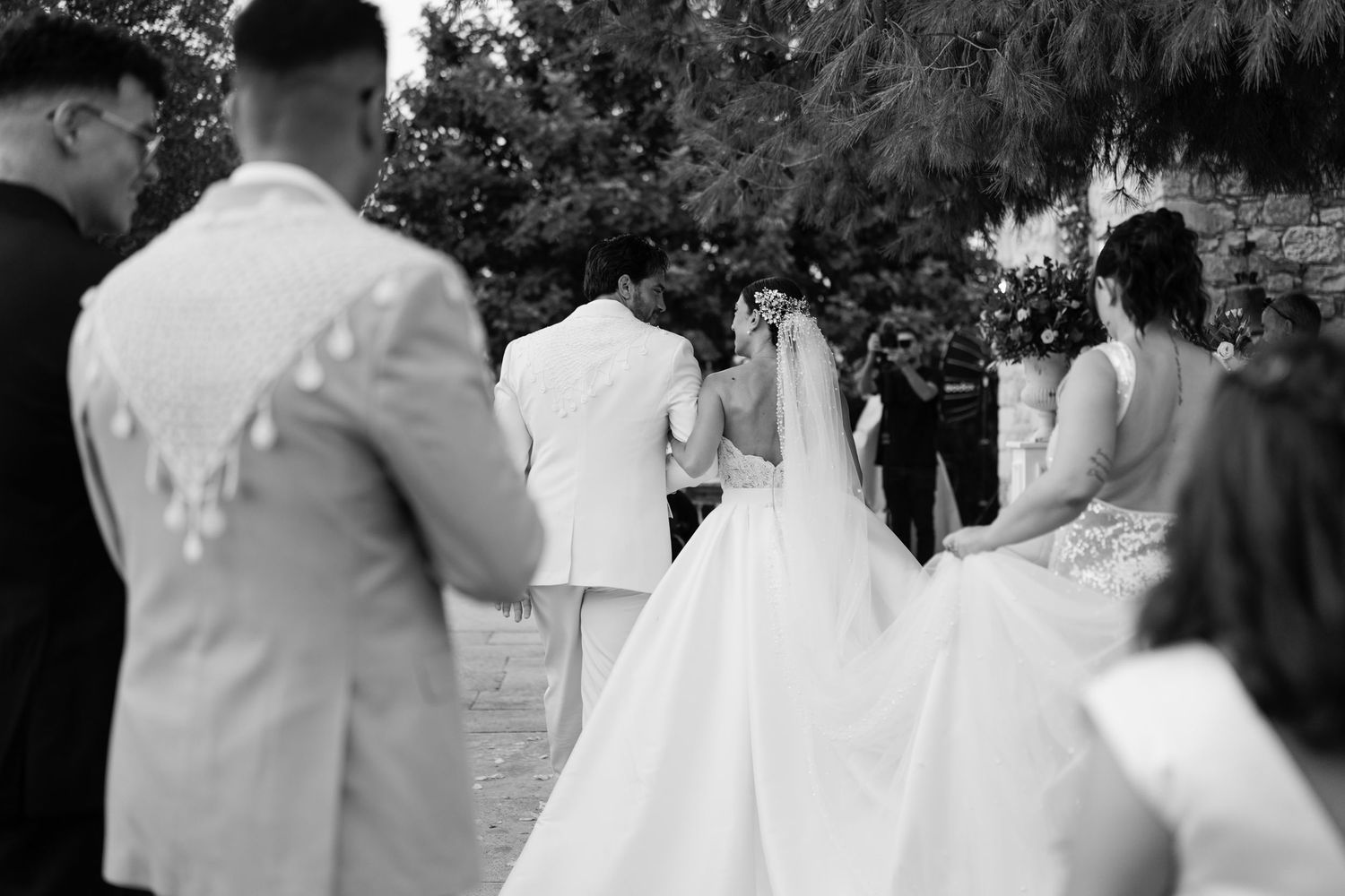 Wedding party celebrates outdoors with joyful embraces and white attire.