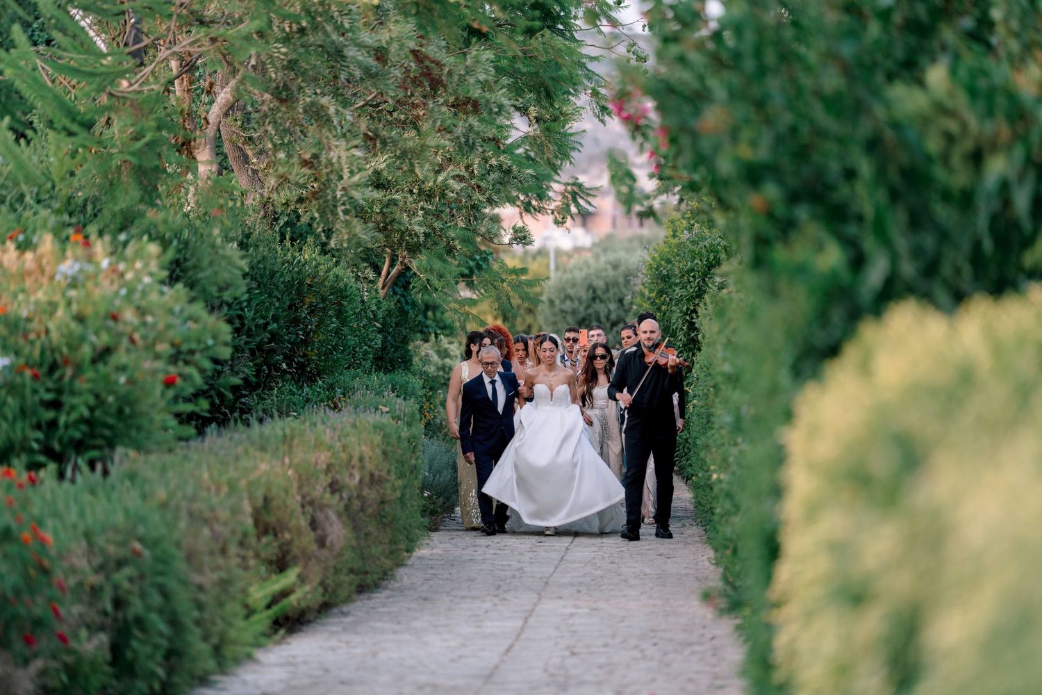 Wedding party walking down tree-lined pathway during outdoor ceremony.