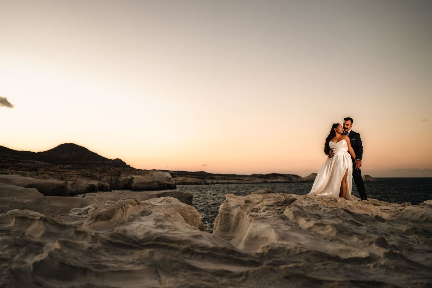 A couple in wedding attire poses dramatically at sunset in a desert landscape.