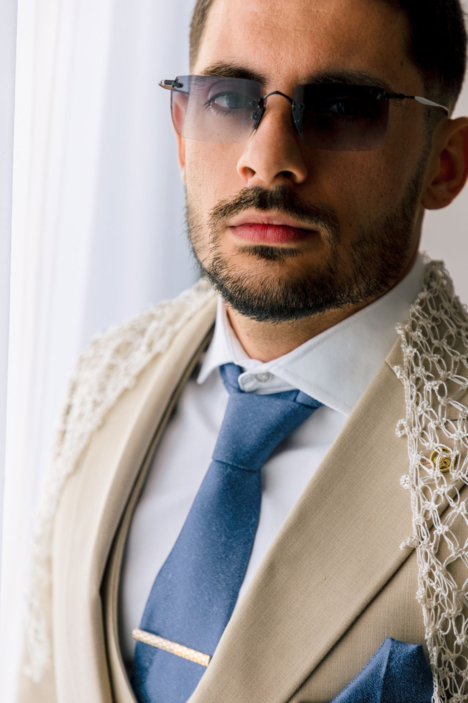 Close-up portrait of a groom wearing a cream suit jacket, blue tie and glasses against a light background.