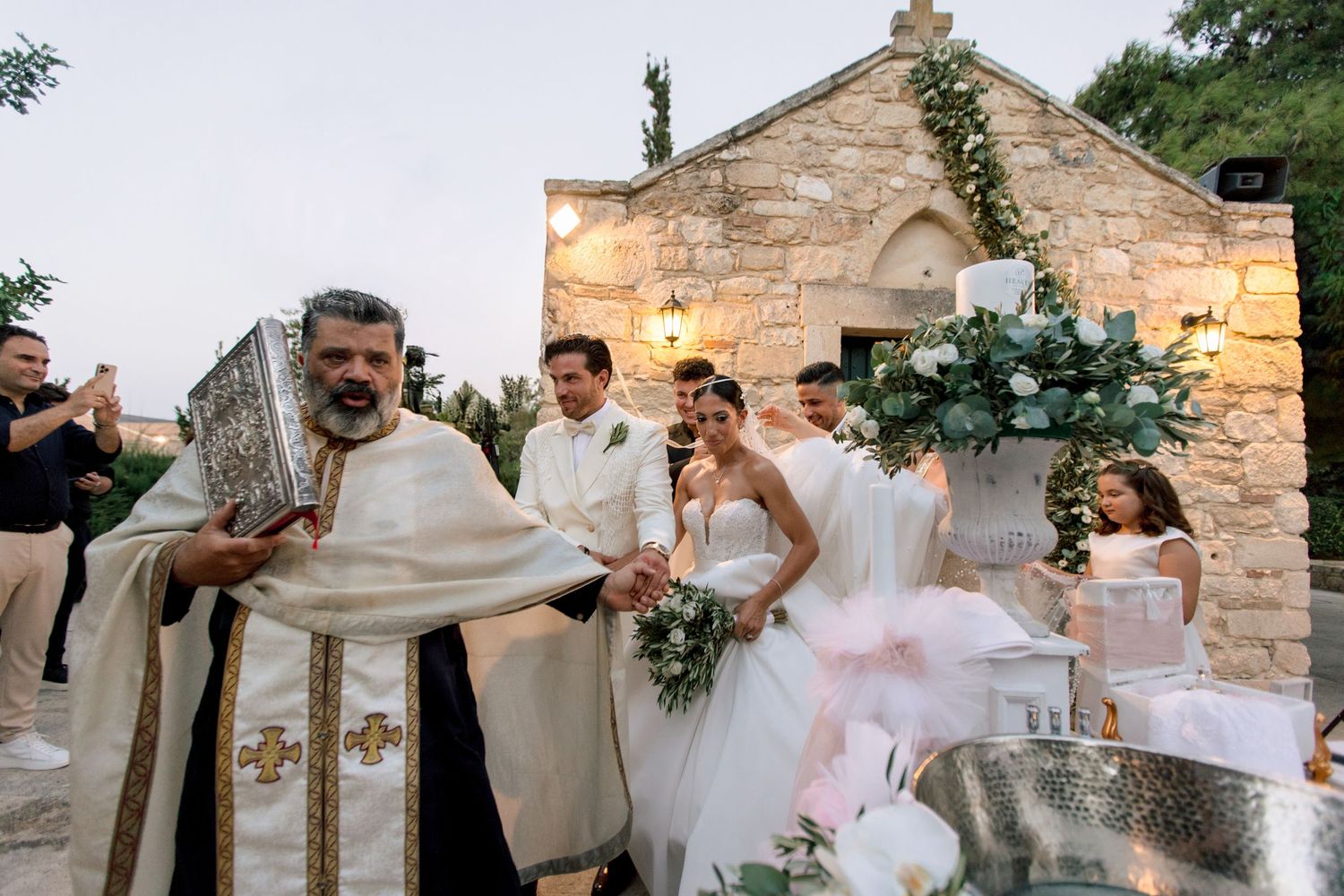 Wedding celebration outside a rustic stone chapel with decorative floral arrangements.