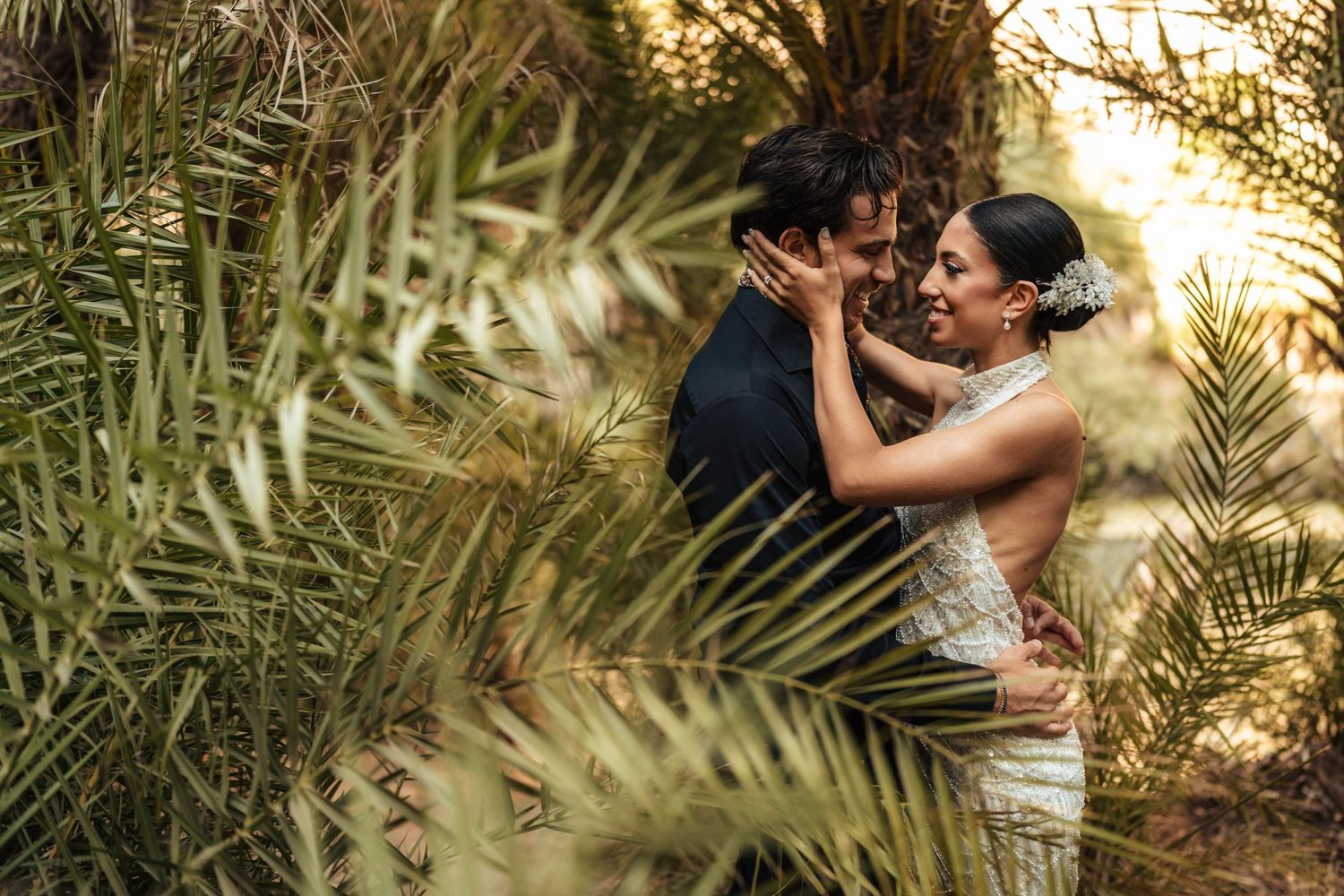 Romantic couple embraces among tropical palm fronds at sunset in an intimate wedding moment.