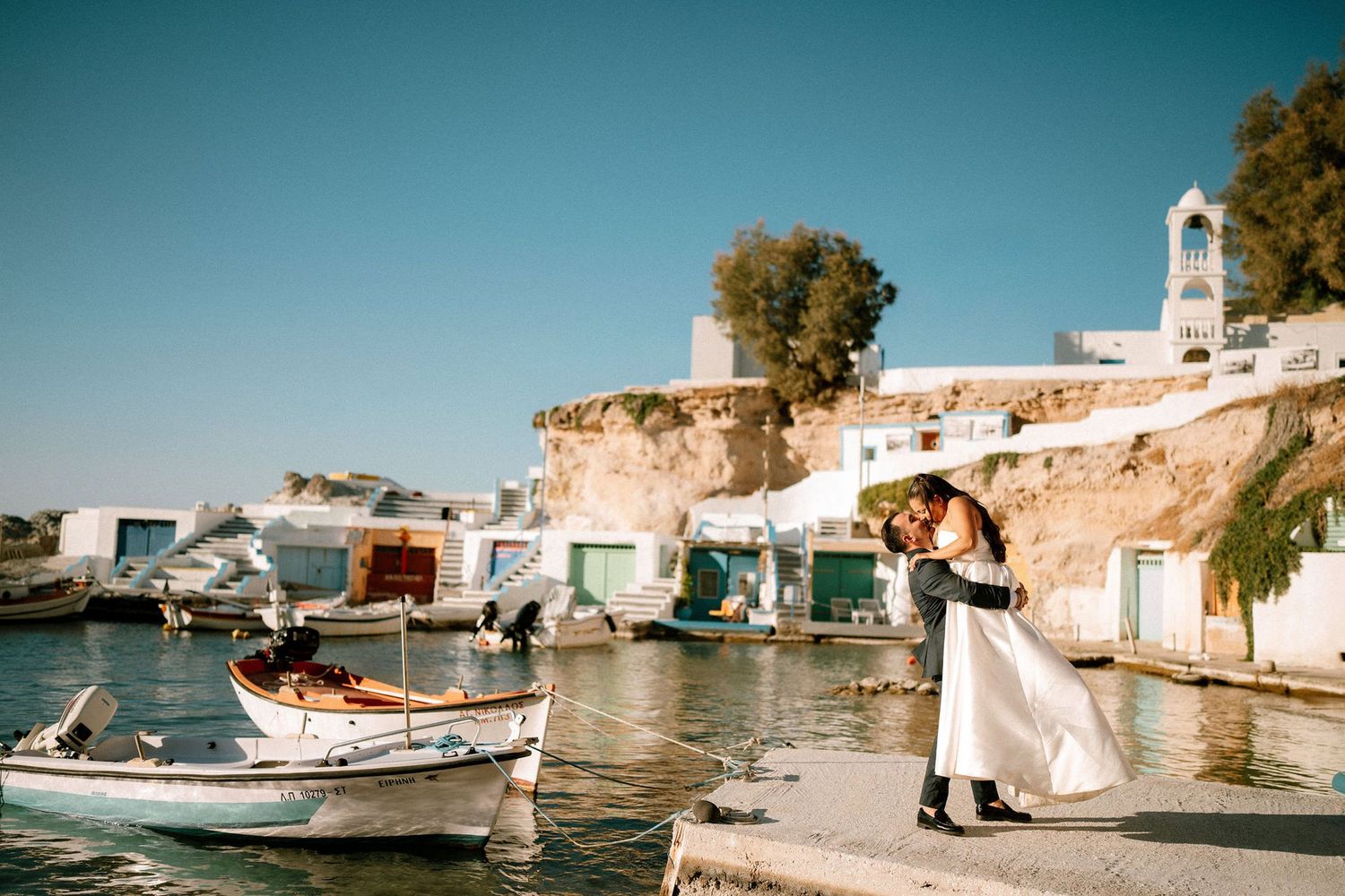 Rustic fishing boats moored along a Mediterranean waterfront with white buildings in the background.