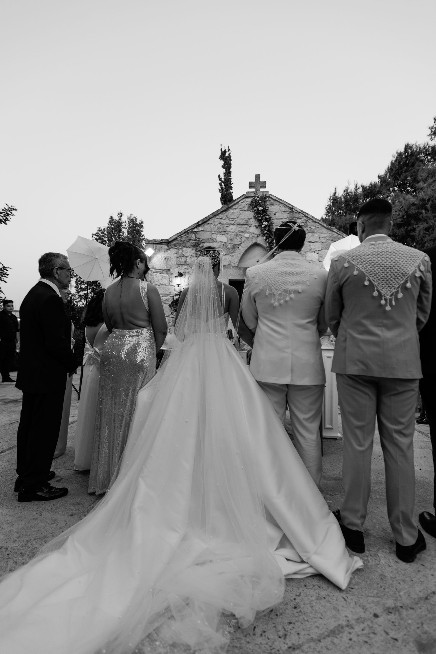 Wedding party members stand together in formal attire against a rustic stone church backdrop.