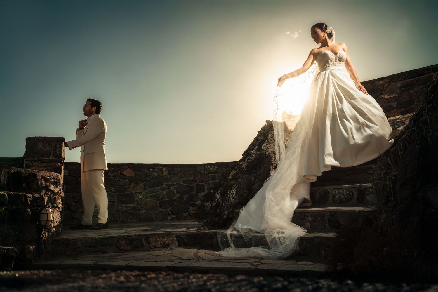 Bride in flowing dress poses dramatically on stone wall at sunset.