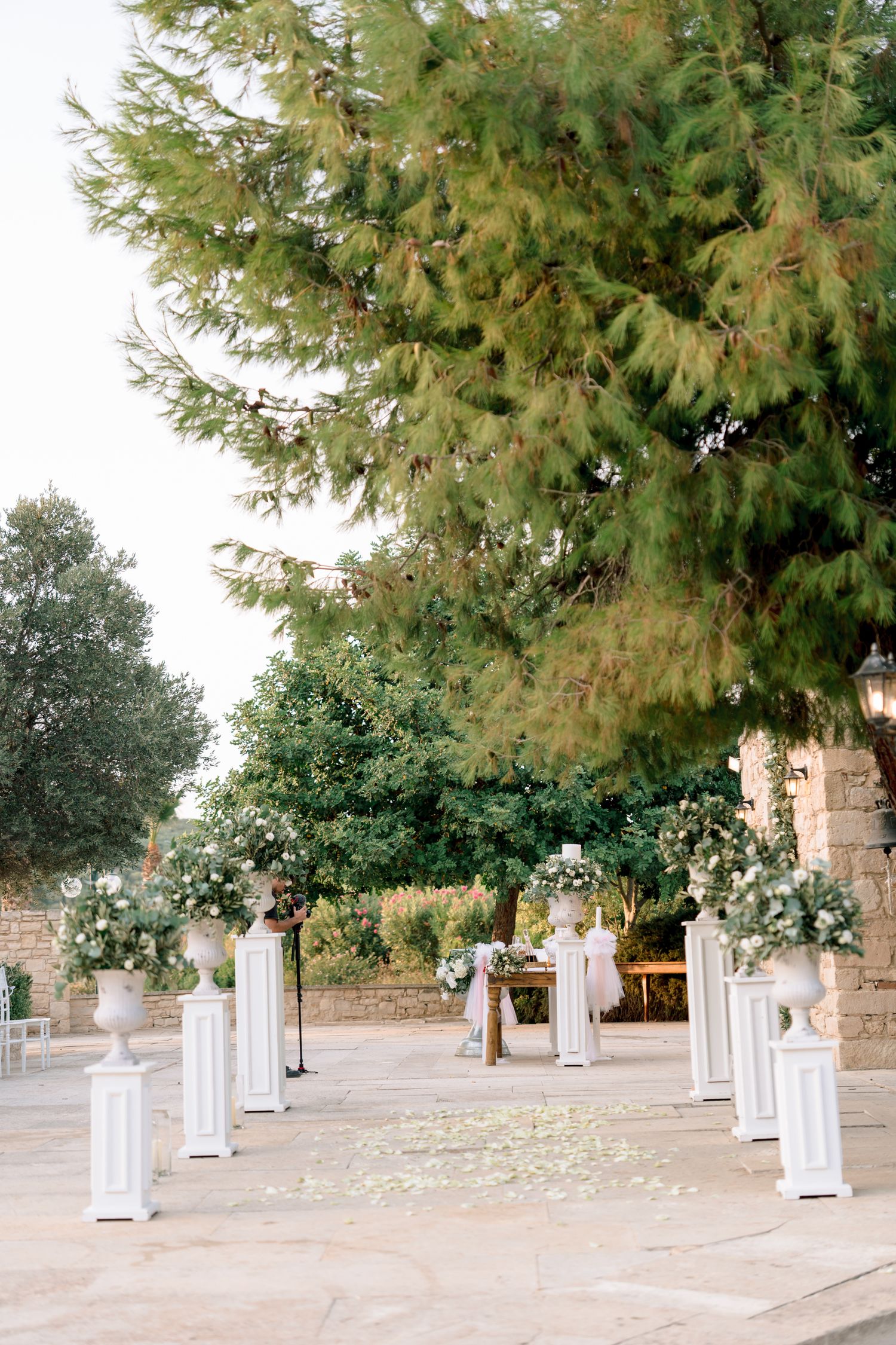 White decorative pillars line a stone pathway under large evergreen trees.