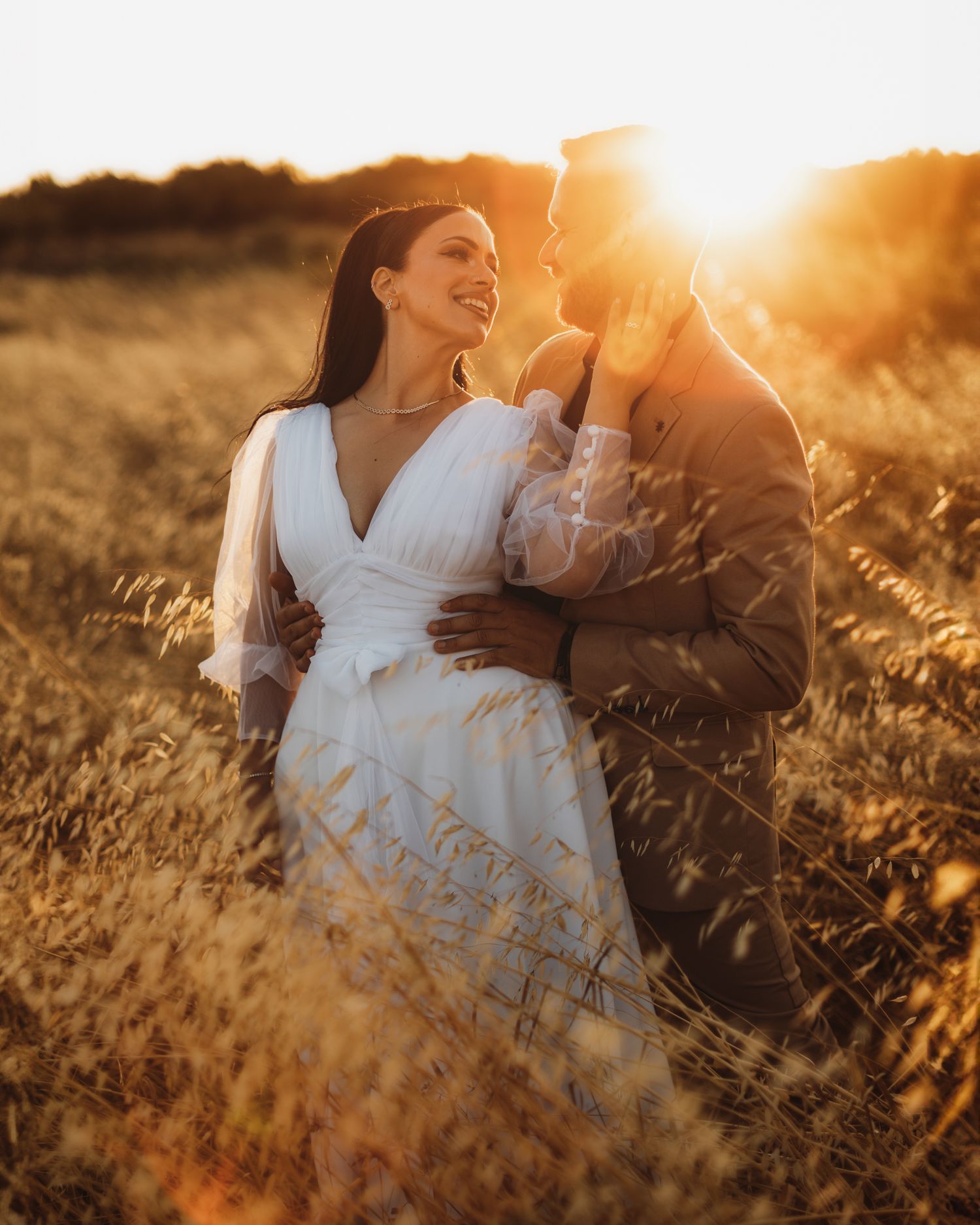 Couple in white clothing sitting among sun-lit wheat fields at golden hour.