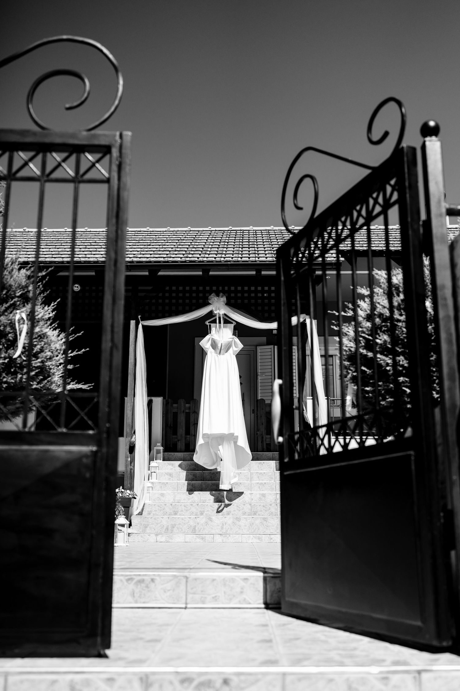 Black and white photo of wedding dress hanging in doorway framed by ornate iron gates.