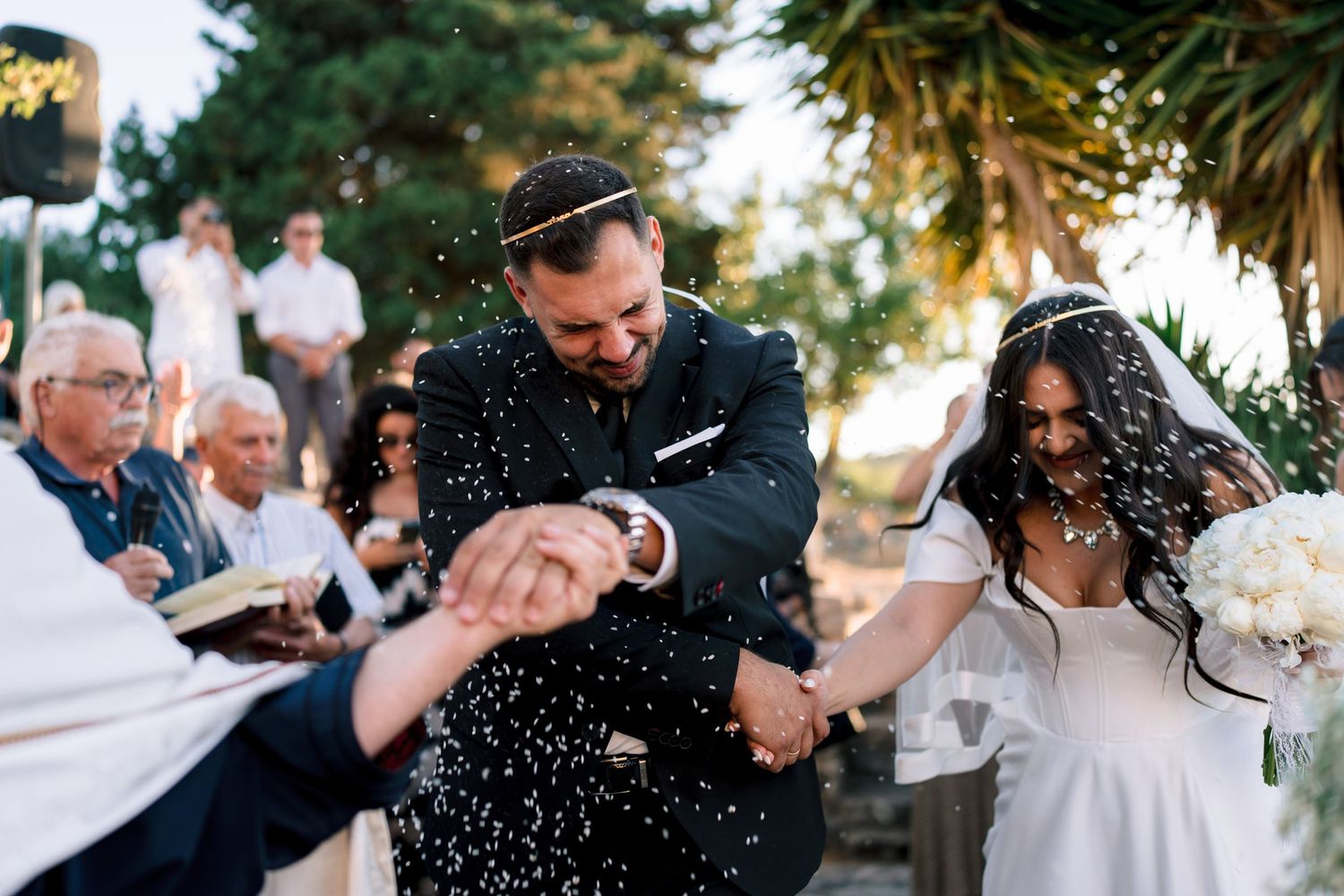 Wedding party celebrates with confetti and laughter in an outdoor garden setting.