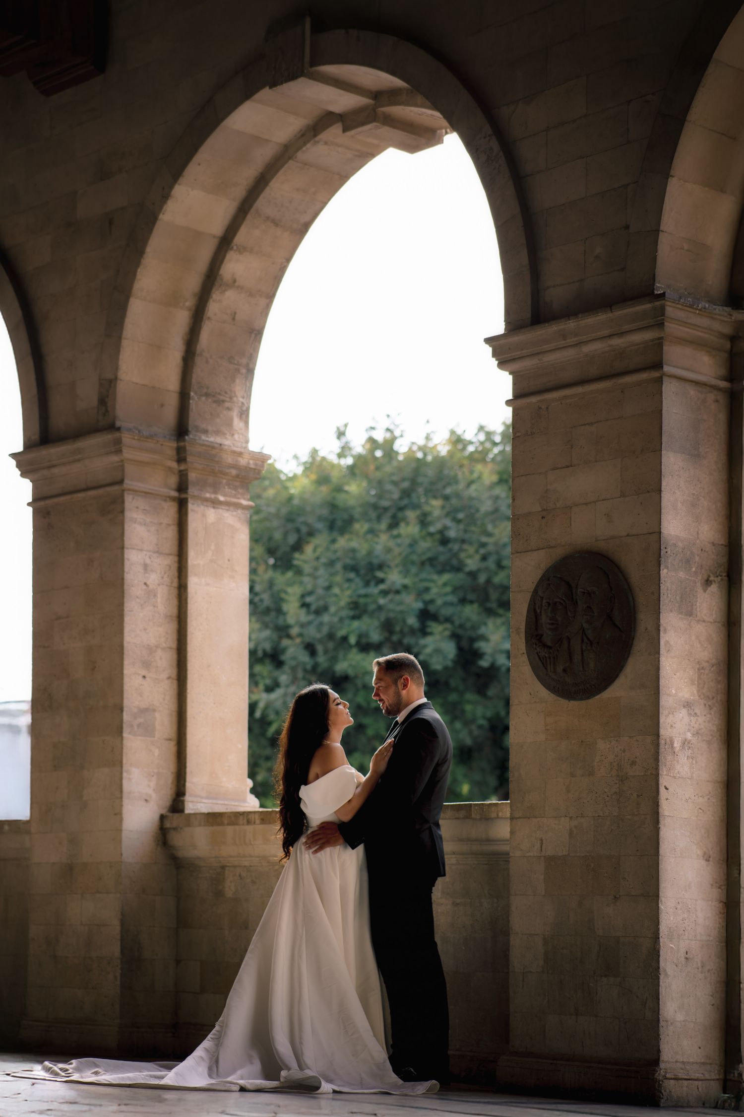 A romantic wedding couple embraces under grand stone archways at sunset.