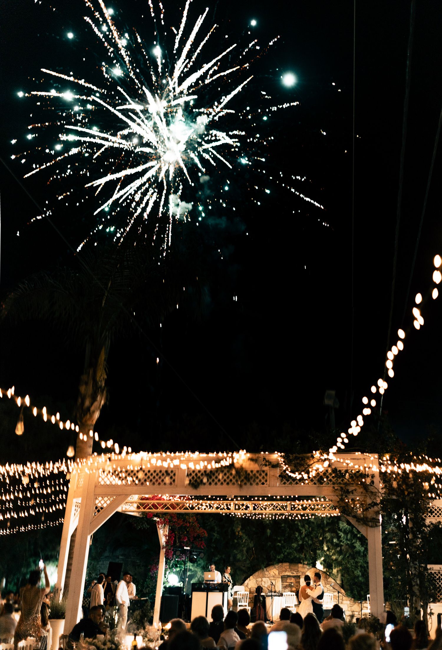 Fireworks illuminate the night sky above a wedding venue decorated with glowing string lights.