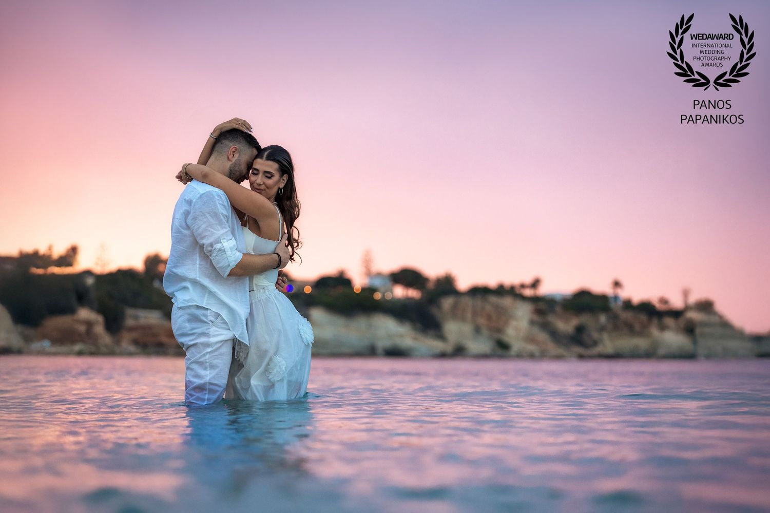 Couple standing together in shallow water during pink sunset for romantic beach wedding photo.