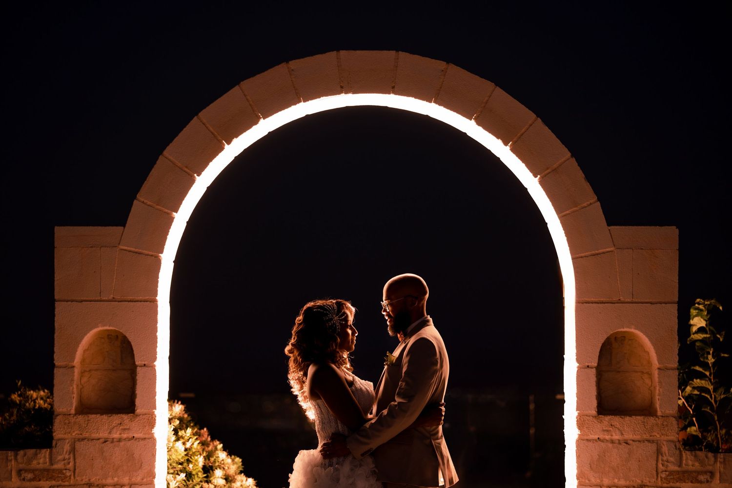 Wedding couple in dramatic backlit silhouette under illuminated stone archway.
