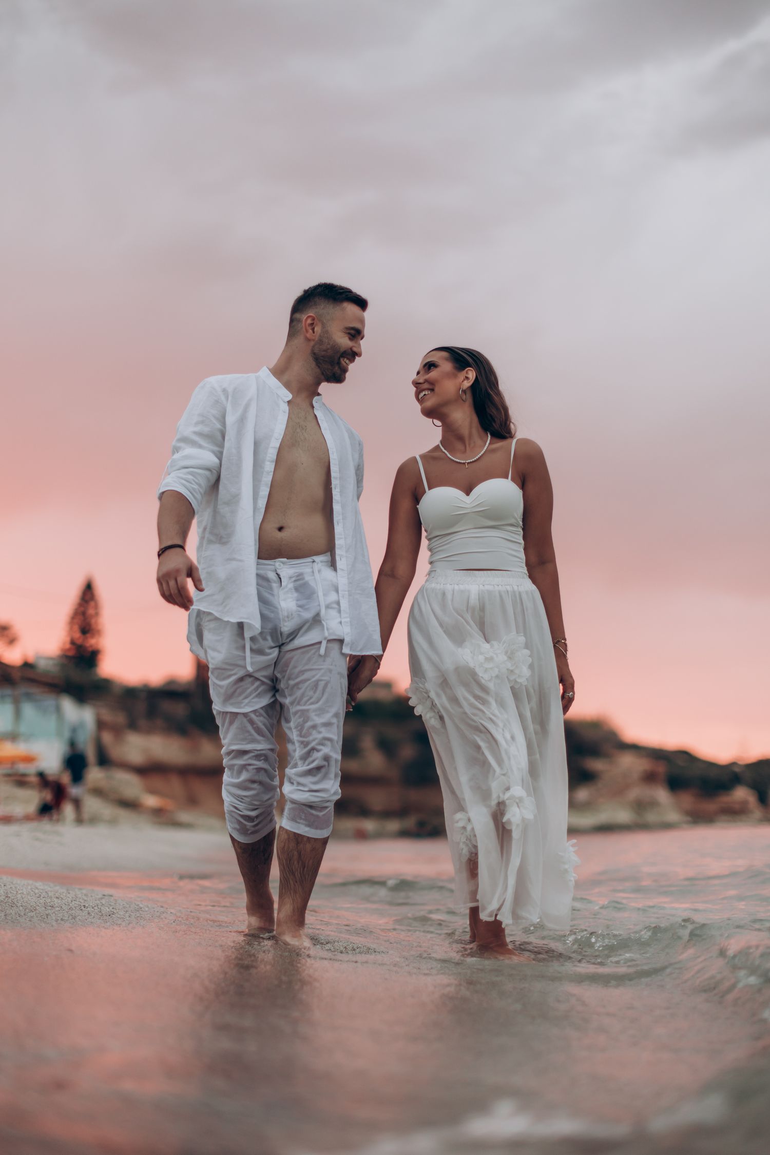 A couple walks along the beach at sunset in elegant white wedding attire.