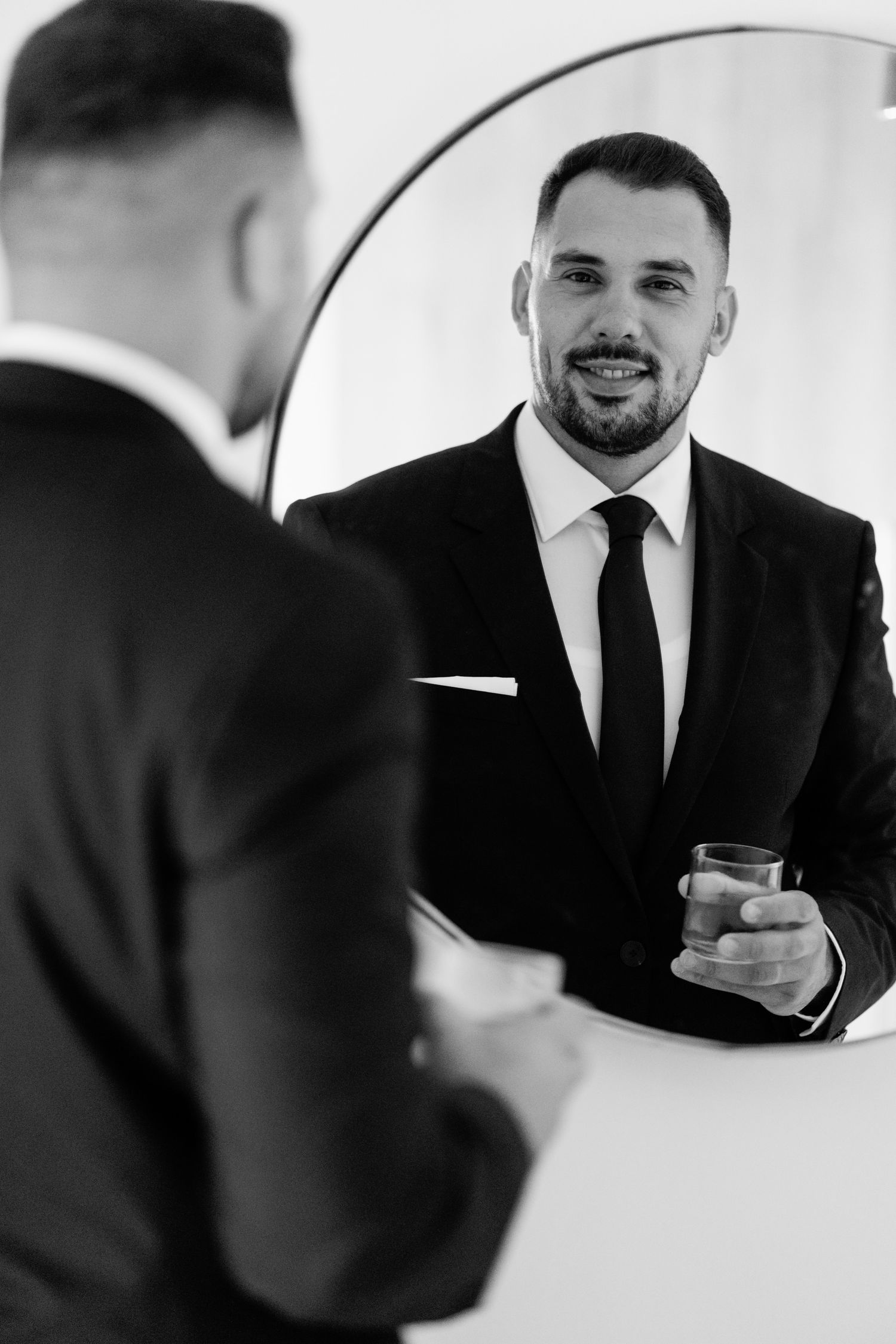 Man in black suit looking in mirror while getting ready.