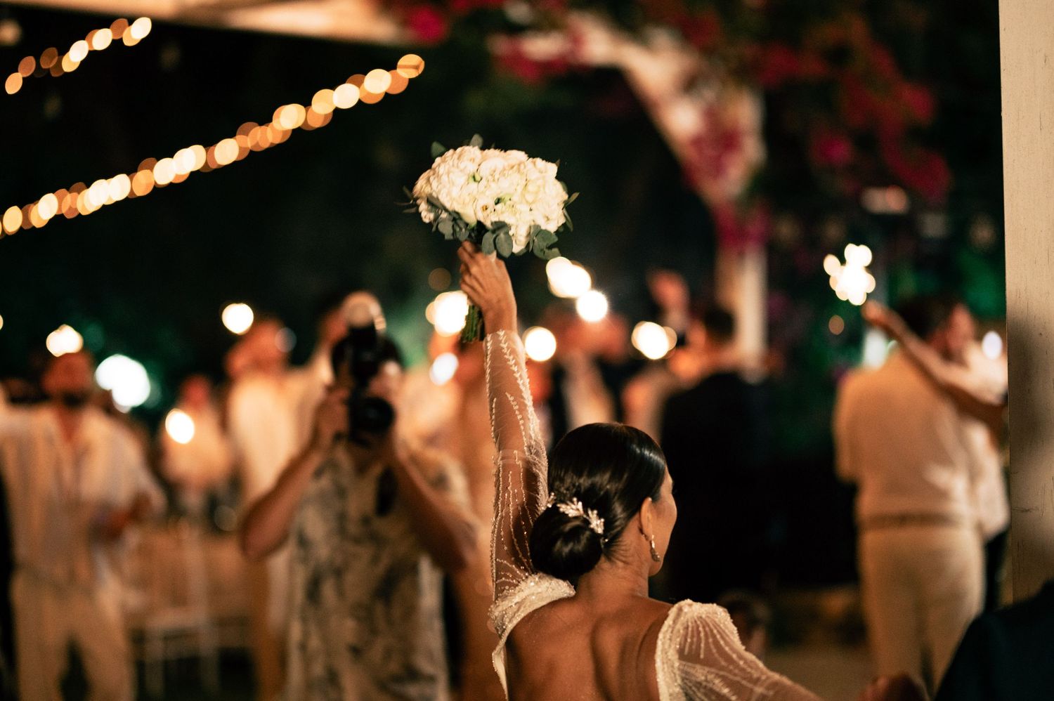Blurred silhouettes of wedding guests dancing under warm string lights at night.