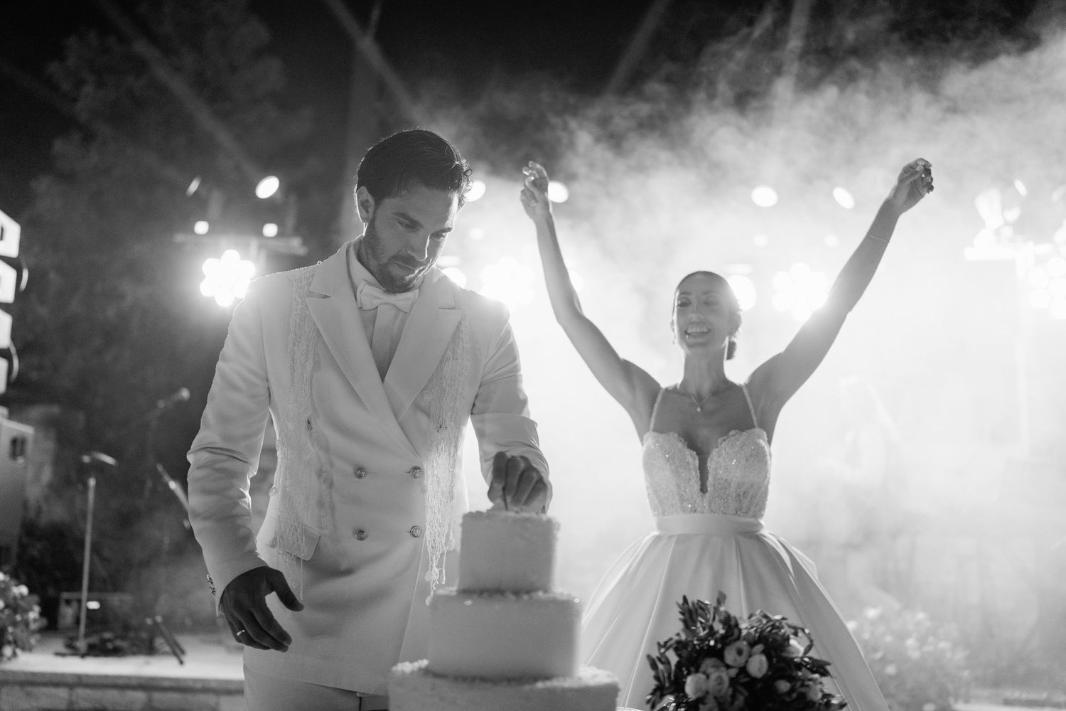 Black and white photo of a couple celebrating by their wedding cake with dramatic backlighting.