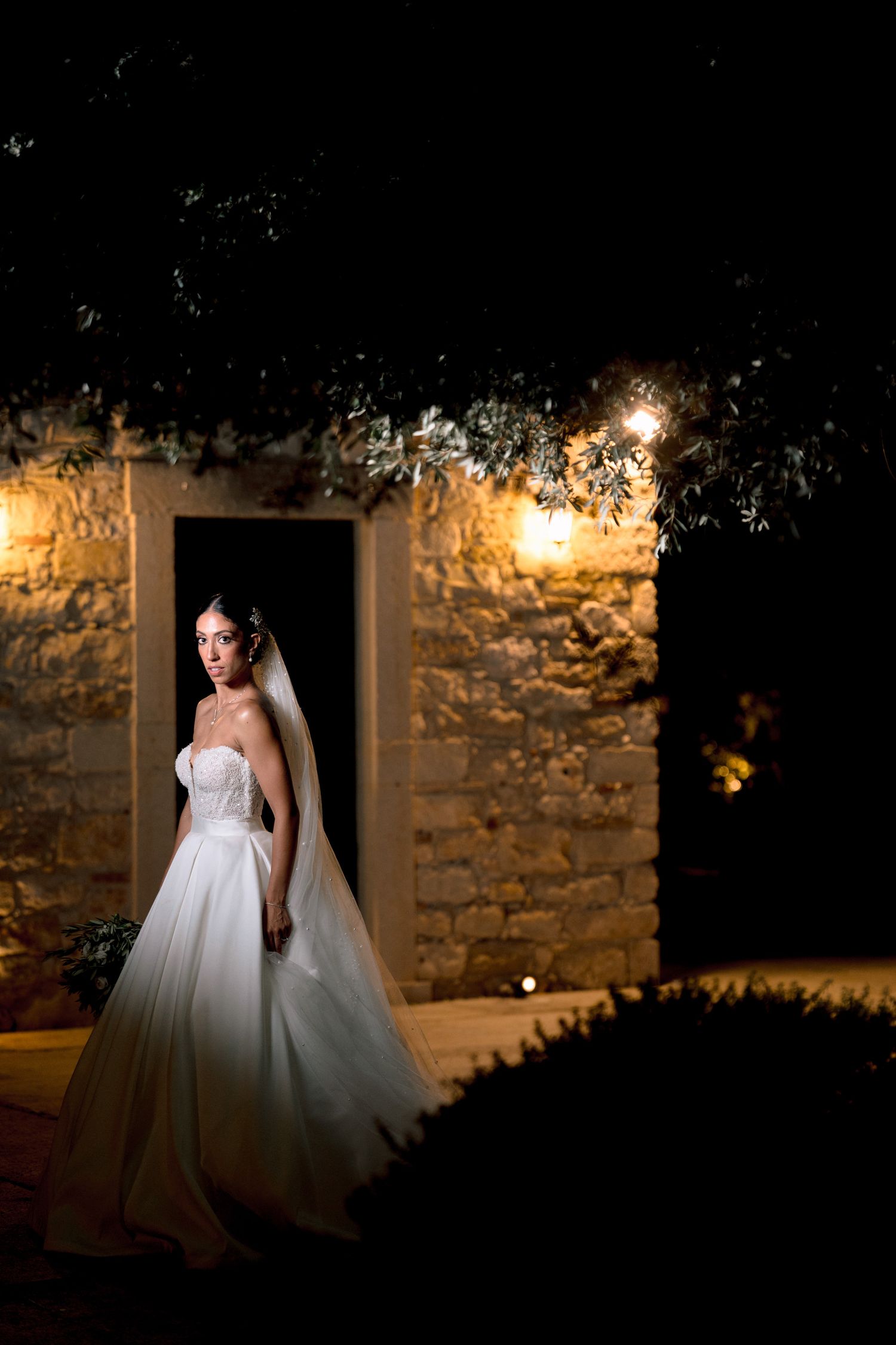 A bride in a white ball gown poses dramatically in a stone doorway at night with warm lighting.