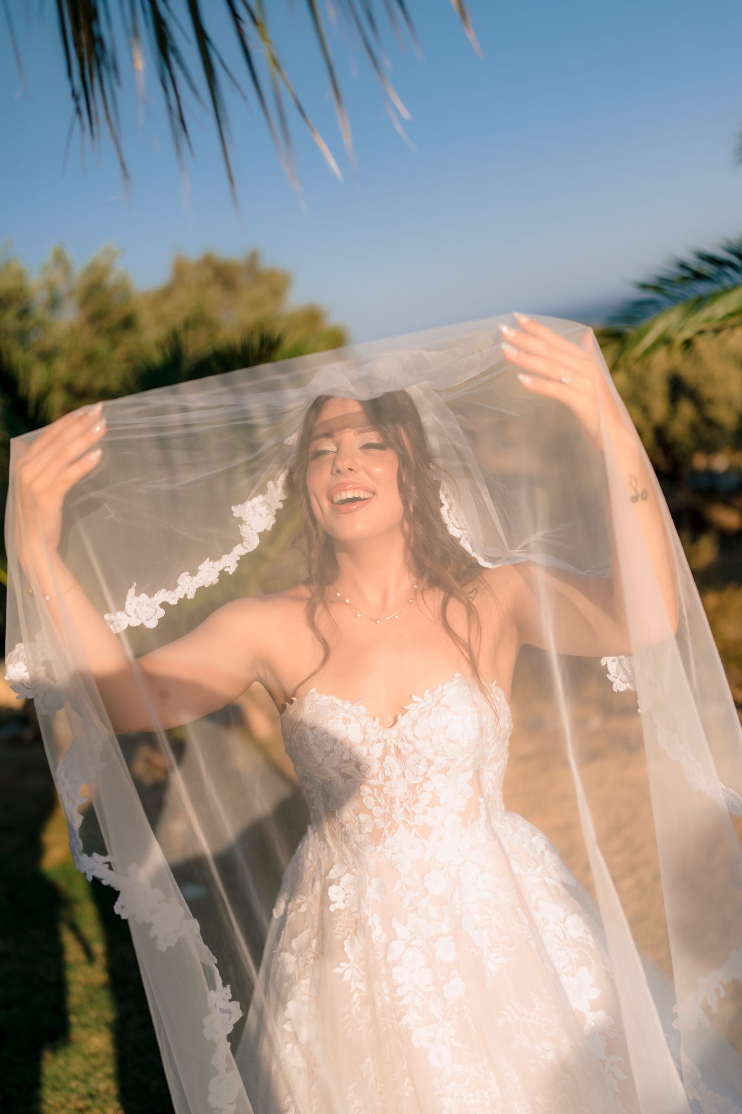 Joyful bride lifts her delicate veil while standing in outdoor tropical garden setting.