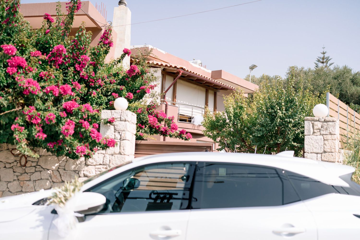 White car parked outside Mediterranean style house with pink bougainvillea flowers.