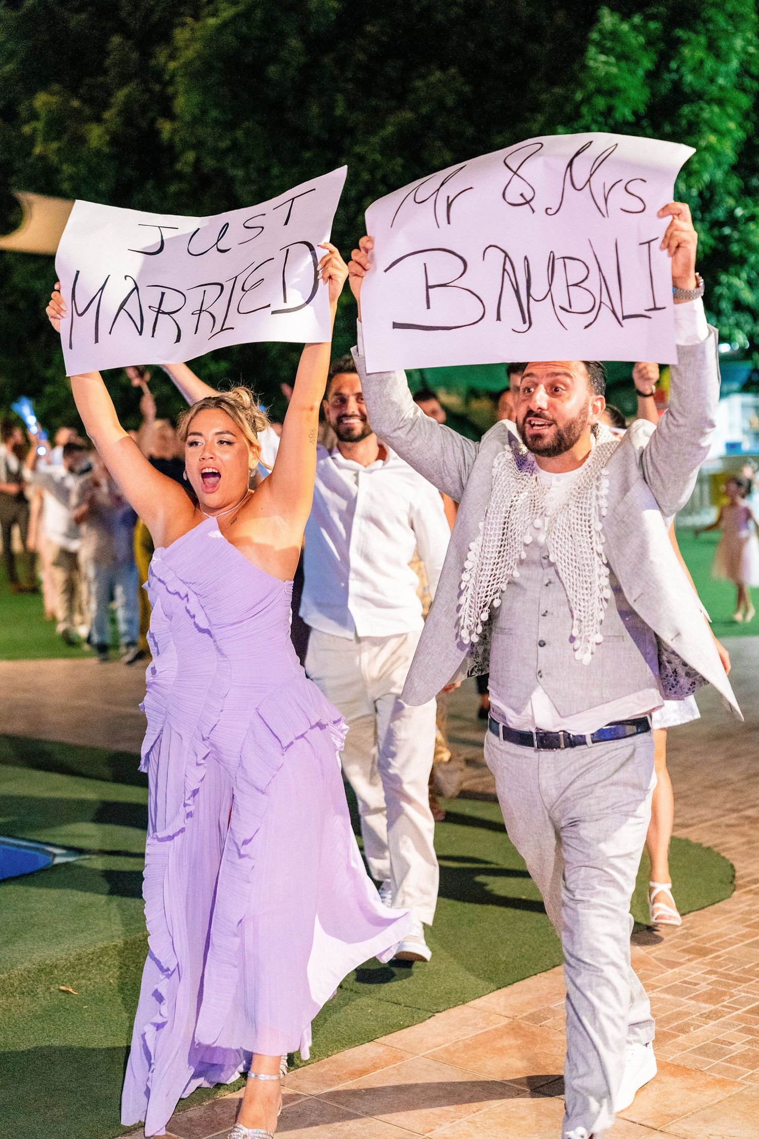 Wedding party in purple dresses and white suits holding celebratory signs outdoors.