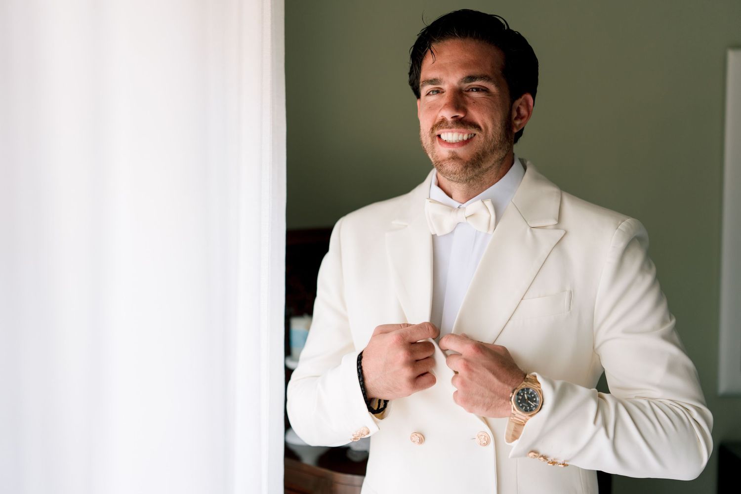 Groom adjusts his white wedding suit while standing near a window in natural light.