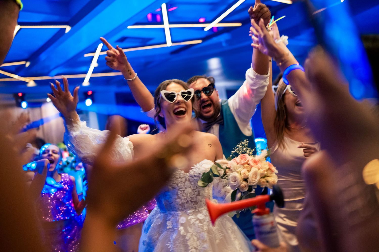 Wedding party dancing under vibrant blue lighting with raised arms in celebration.