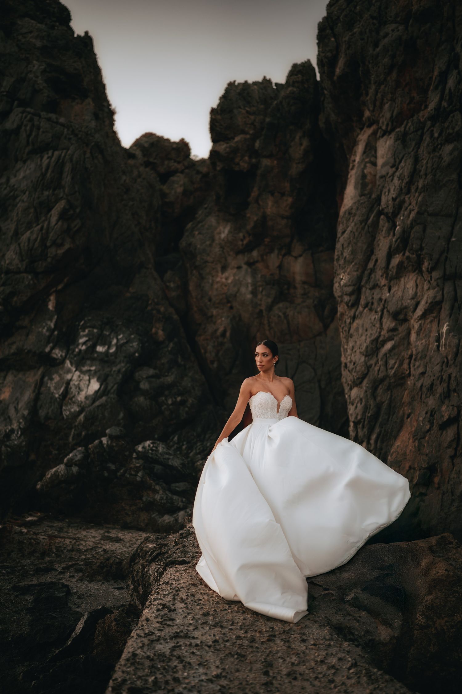 A bride in a flowing white gown poses dramatically against rocky cliffs.