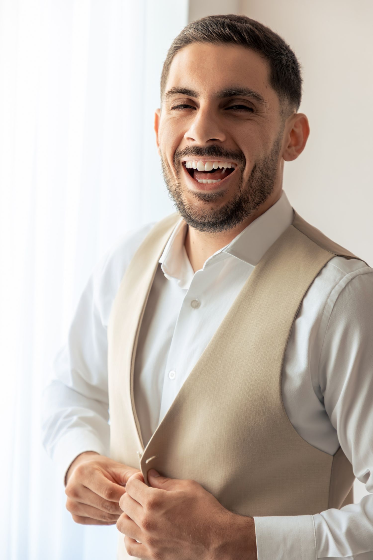 A groom in a white shirt and beige vest smiles warmly while preparing for his wedding day.