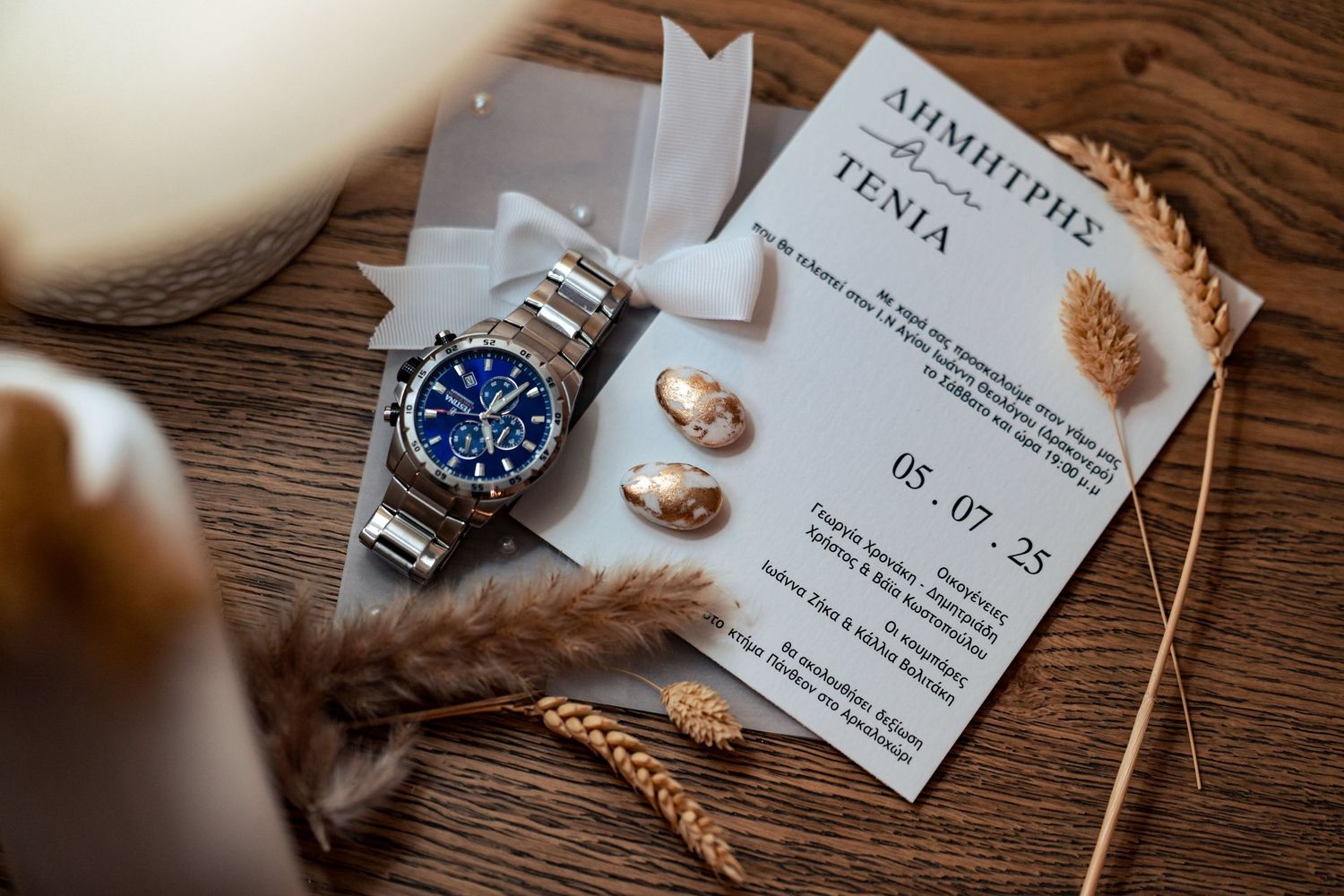 Wedding details flatlay with blue watch, rings, and invitation on wooden surface.